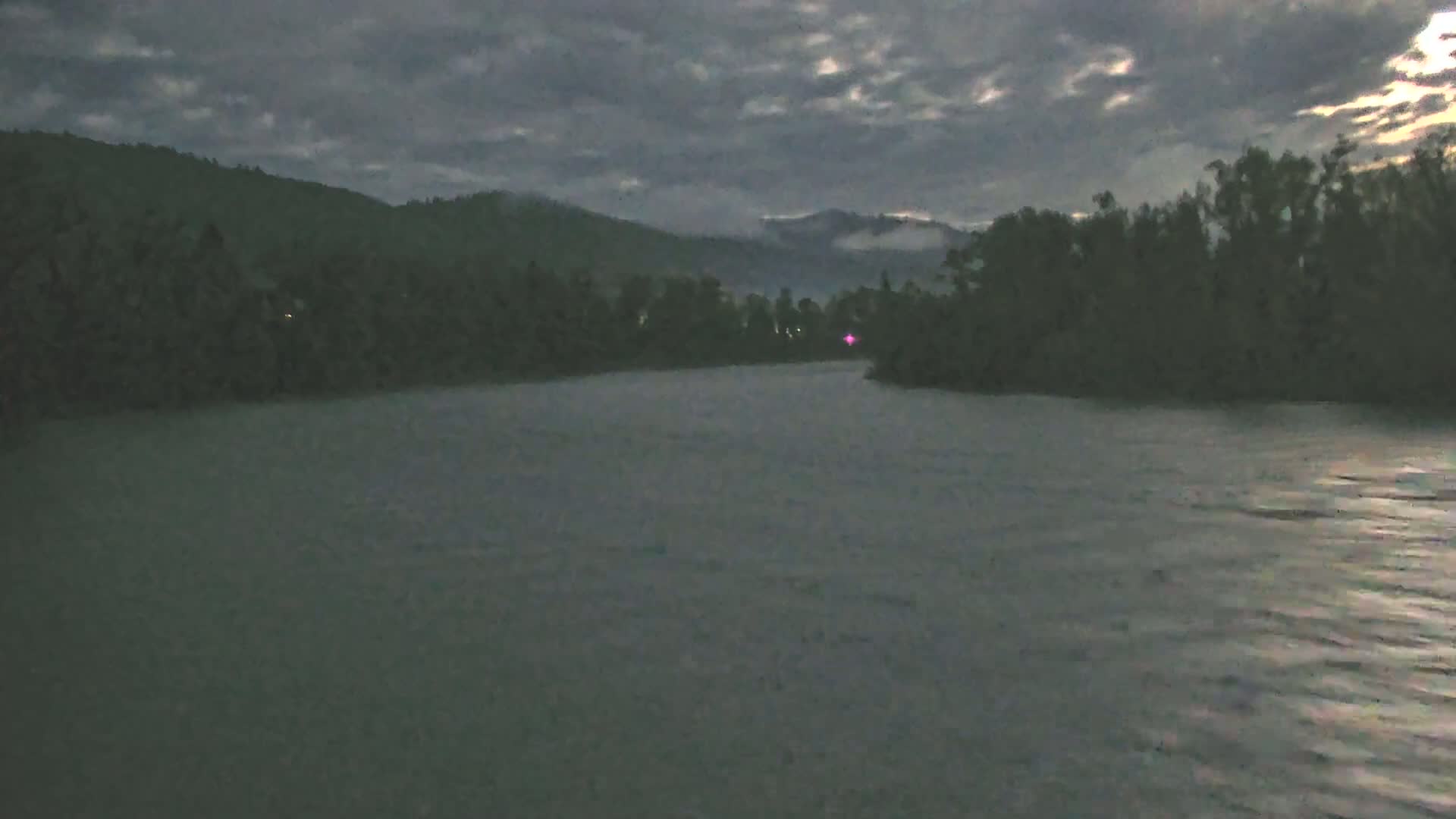 A dark, cloudy evening scene shows a calm river flowing past a tree-lined bank and into the background of dark, distant mountains.