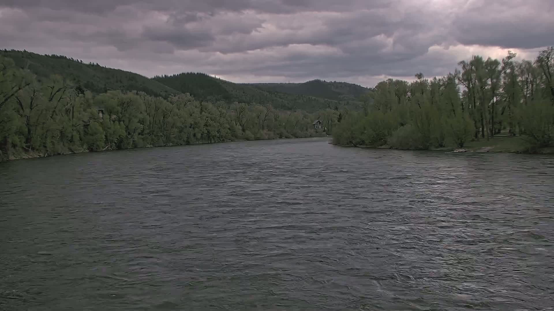 A wide river flows under a cloudy sky, past lush green banks lined with trees and a distant house.