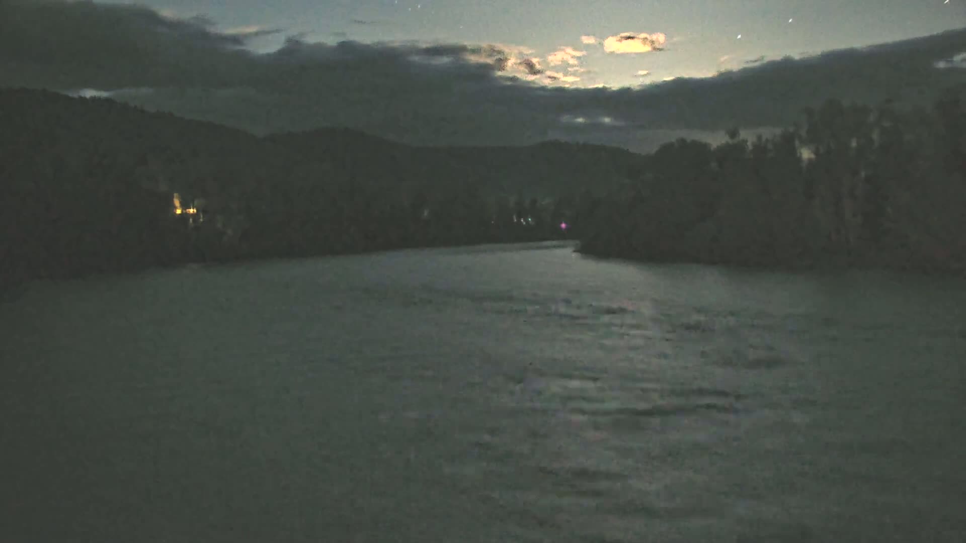 A dark, moonlit night scene shows a calm river flowing between forested hills under a partly cloudy sky.
