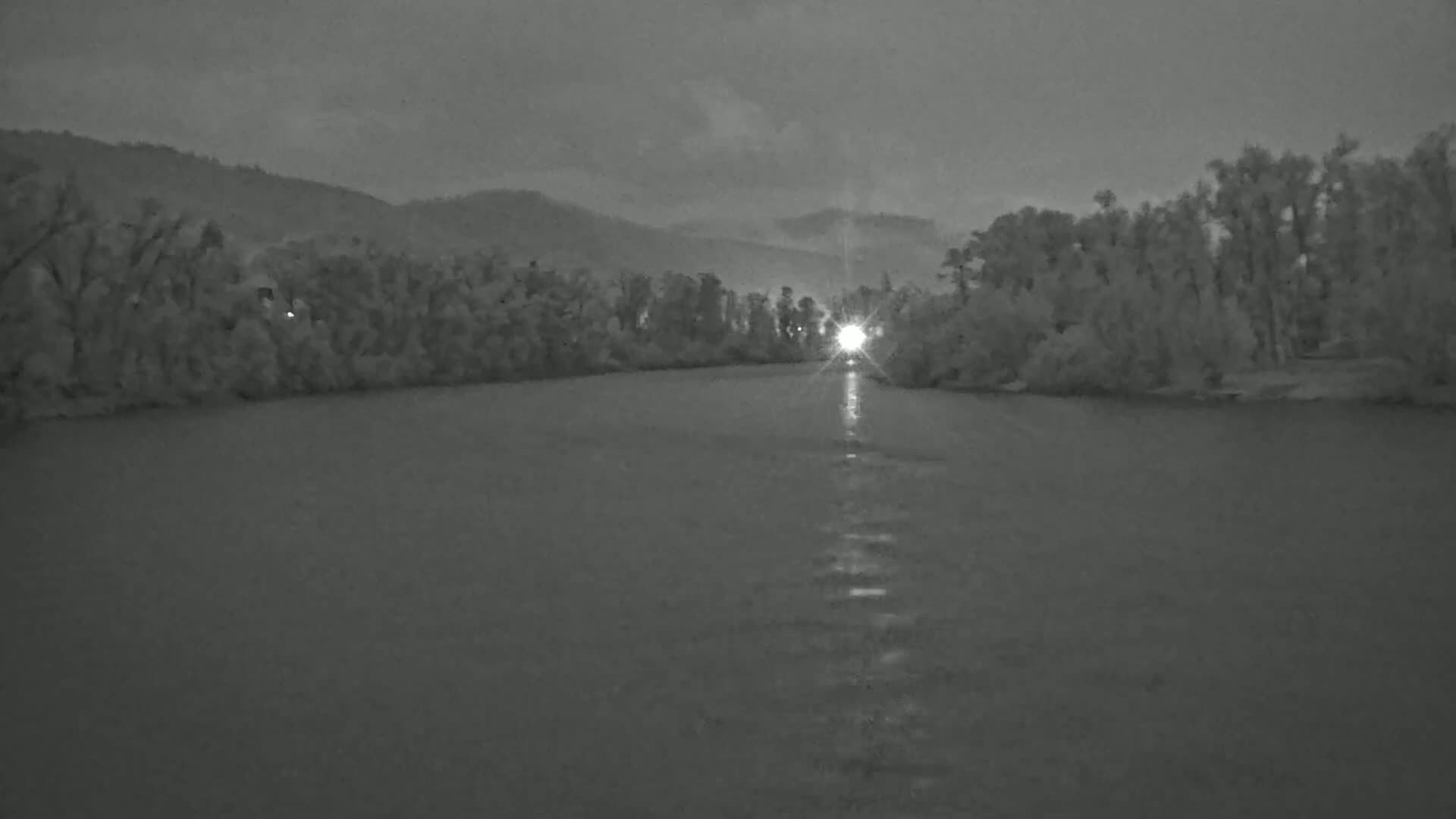 A bright light shines on a calm river that runs between tree-lined banks beneath an overcast sky.