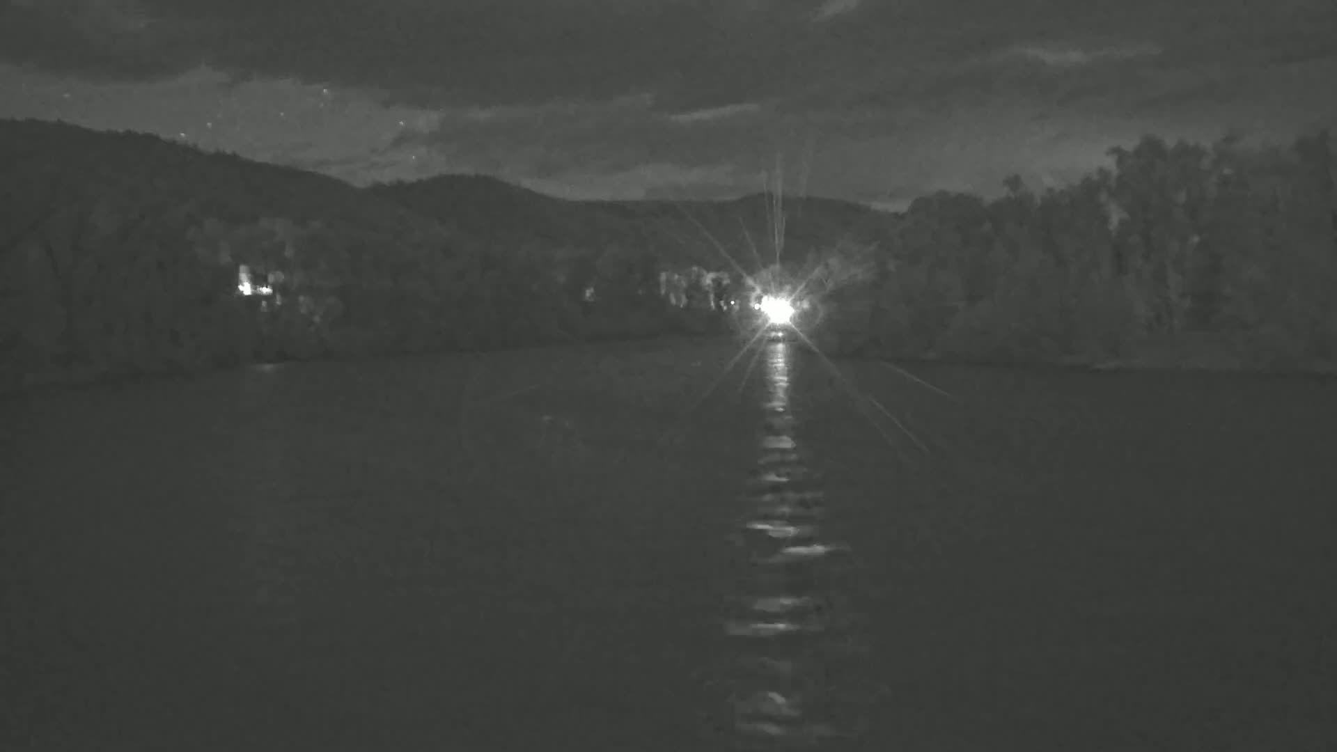 A bright light on a body of water at night is reflected on the dark, calm water, with trees and hills in the background under a partly cloudy sky.