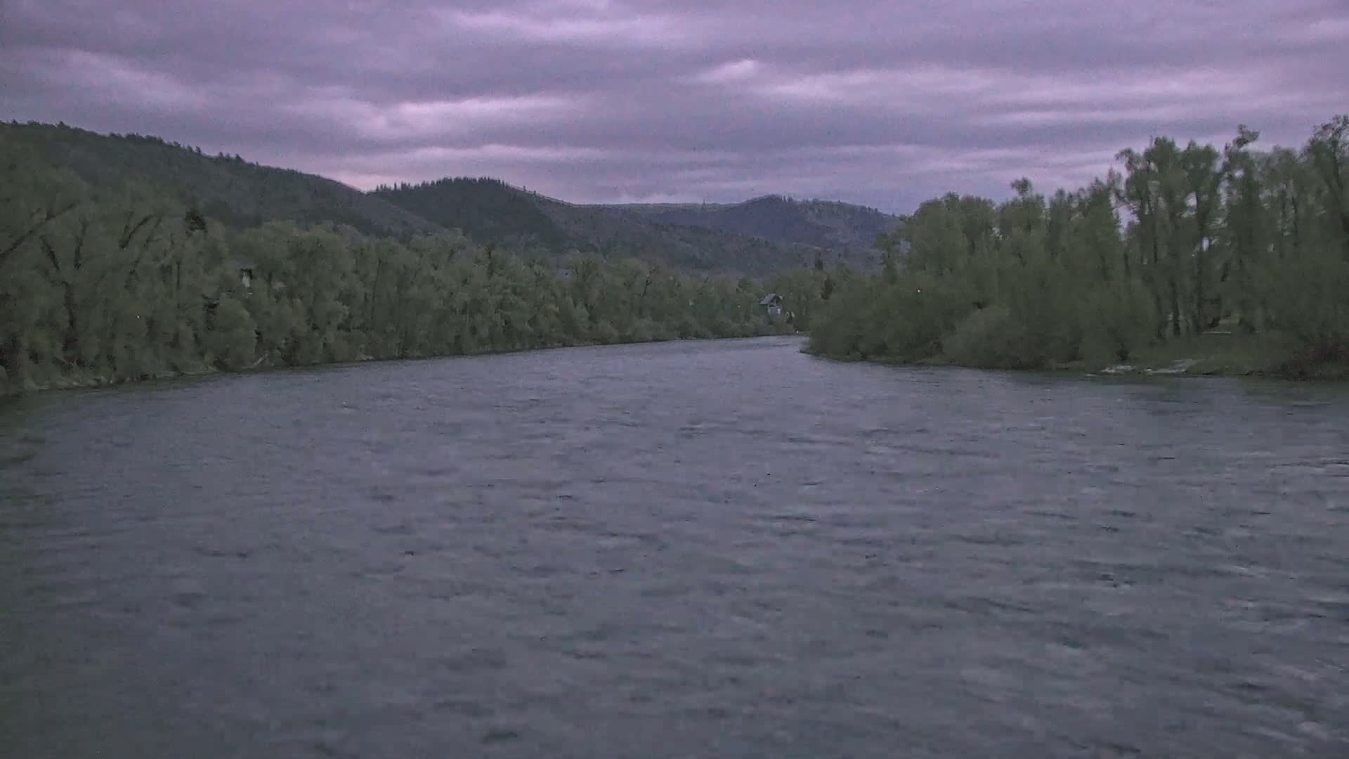 A wide, gently flowing river under a cloudy, dusky sky is surrounded by lush green trees and hills.