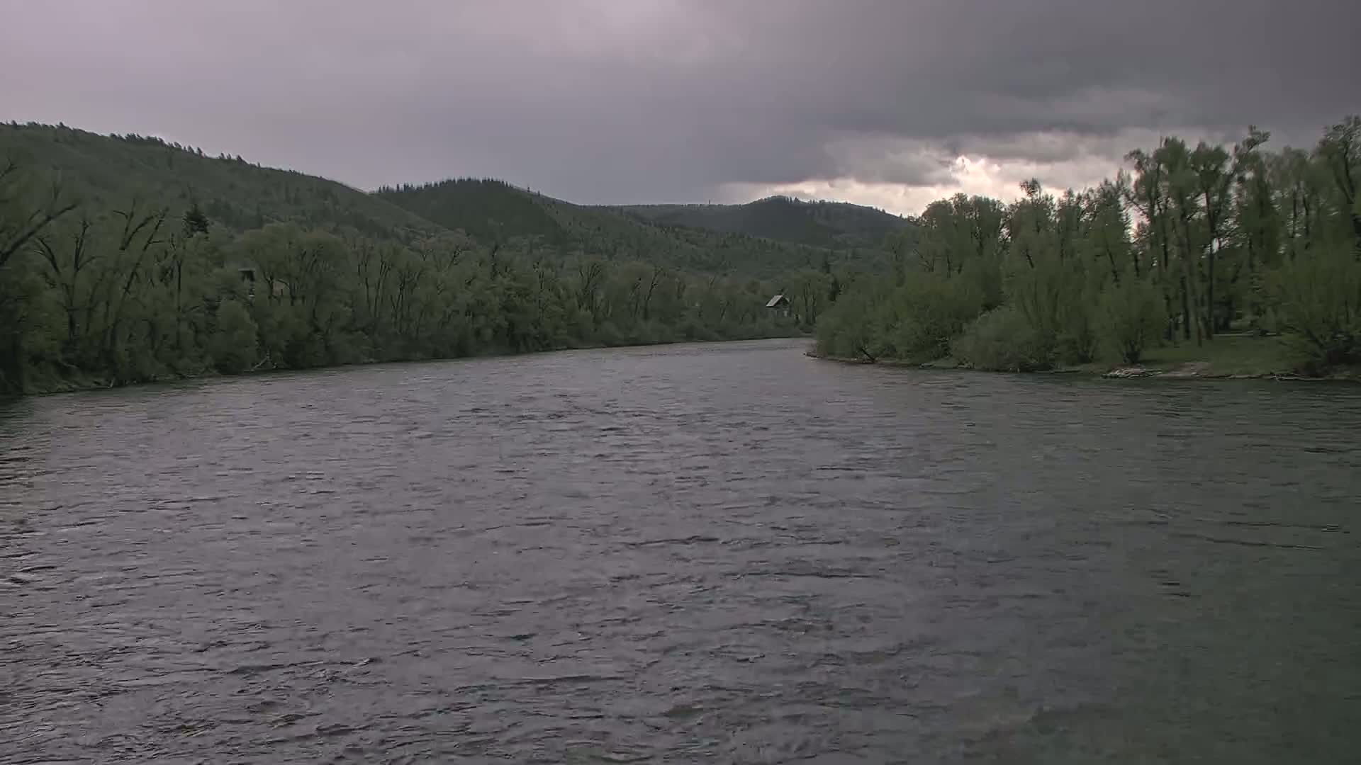 A wide, dark river flows under a gray, cloudy sky past lush green hillsides lined with trees.
