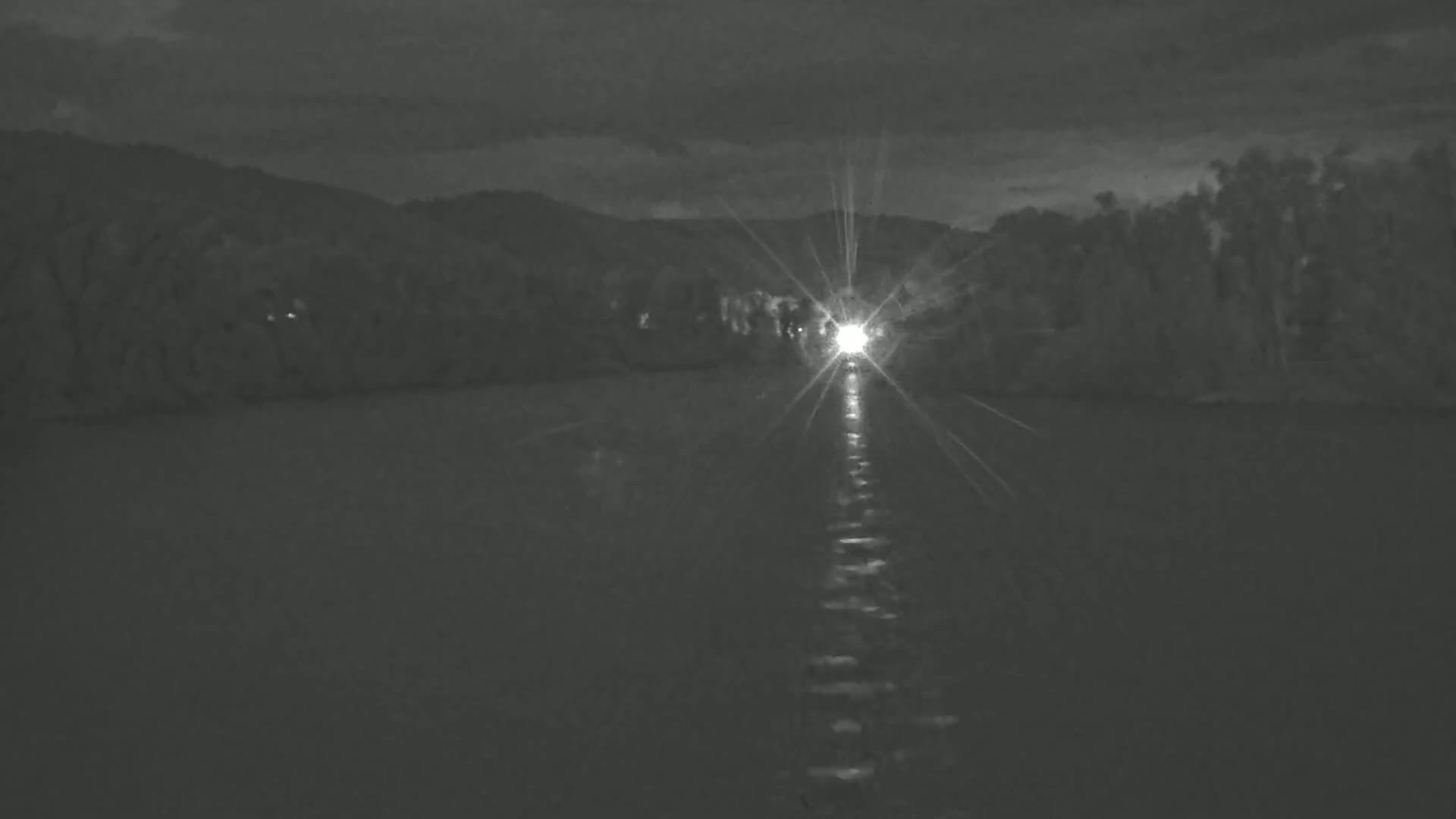 Headlights from a vehicle approaching on a dark, calm body of water, reflecting off the surface under a cloudy night sky.