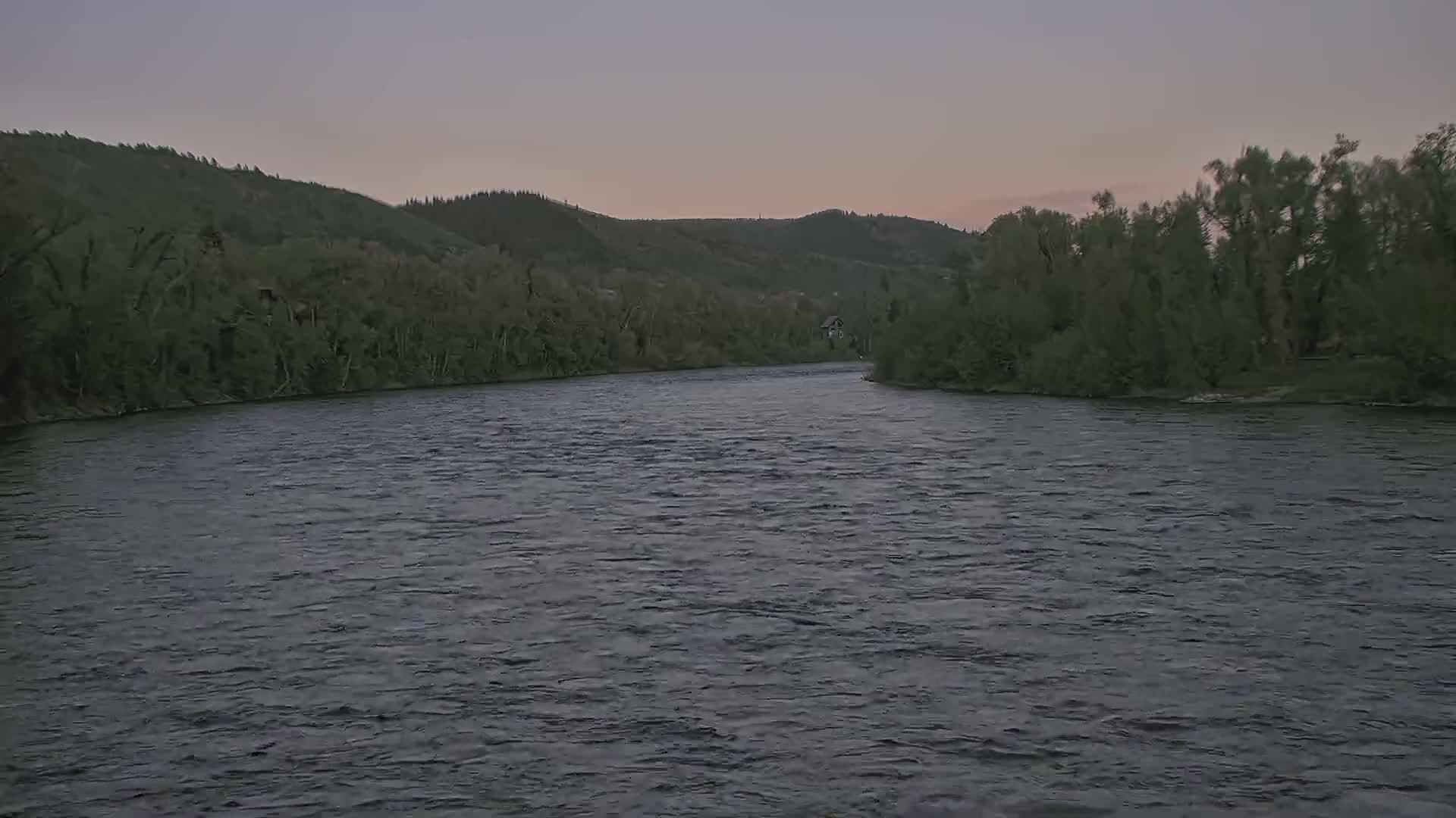 A wide, gently flowing river curves between banks lined with dark green trees under a dusky sky.