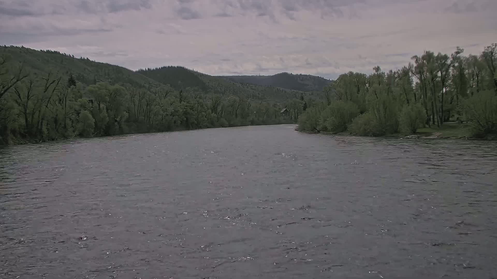 A wide, grey river flows under a cloudy sky, bordered by lush green trees and hills.
