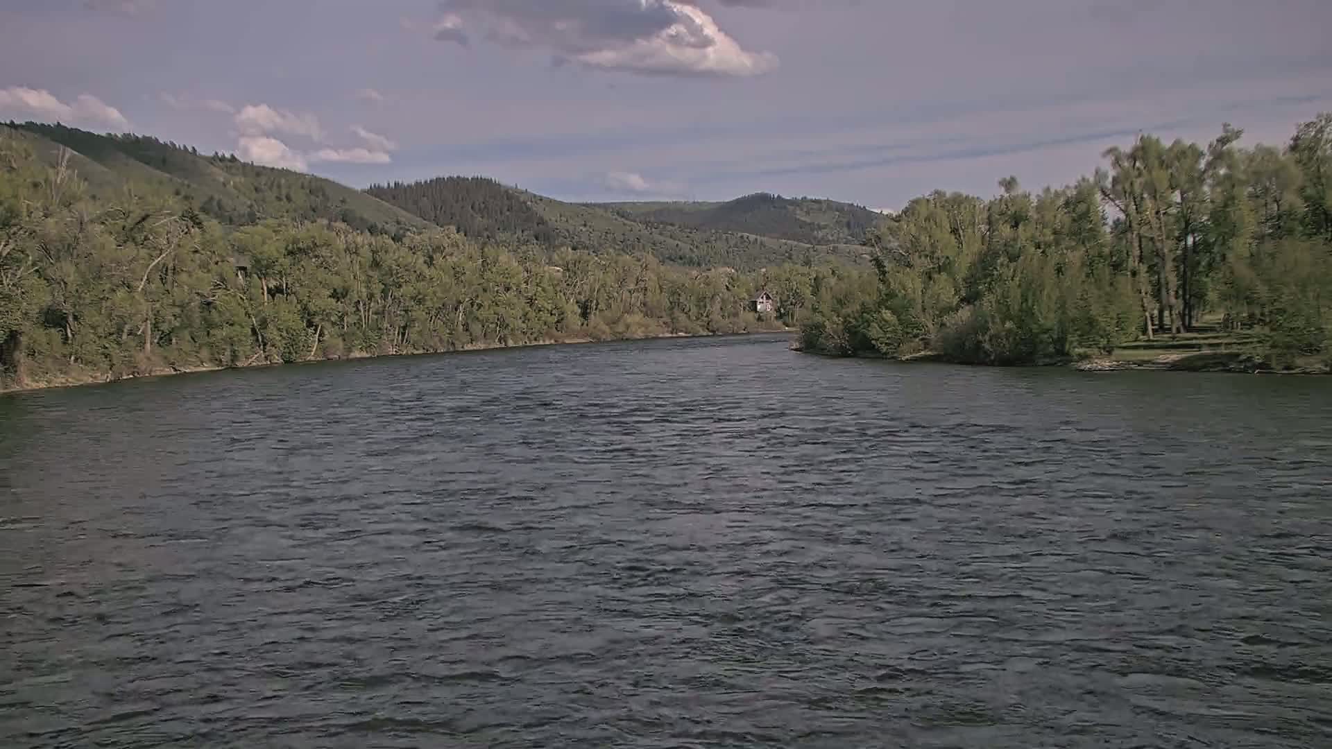 A wide, dark river flows between lush green banks under a partly cloudy sky.