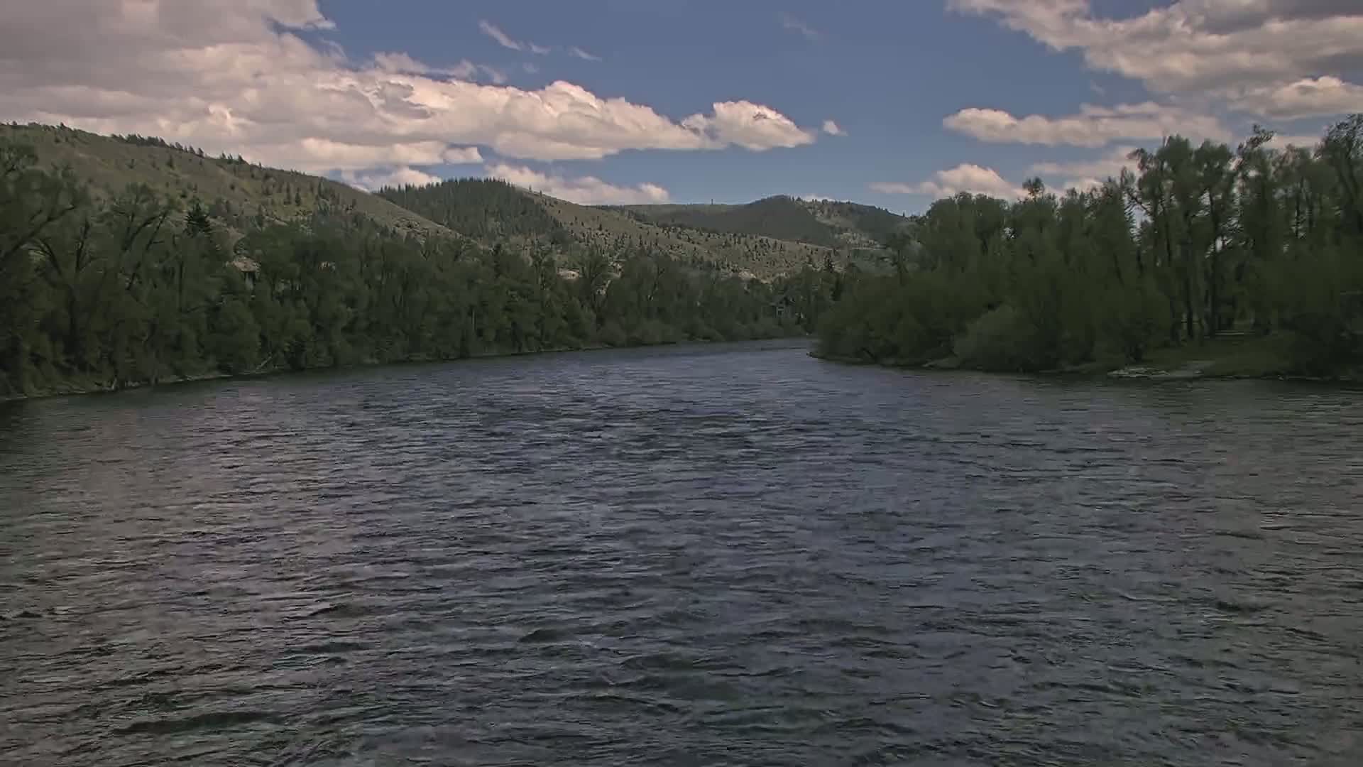 A wide, dark river flows under a partly cloudy sky past verdant, tree-lined banks and hills.