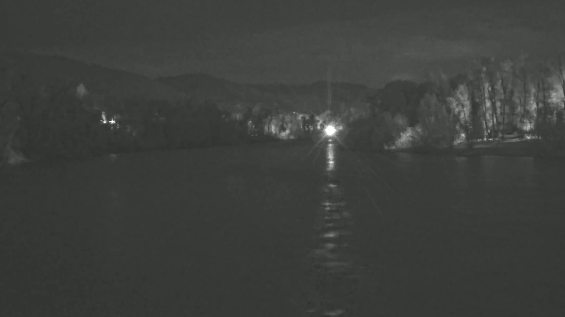A nighttime view of a dark river reflecting the lights of a vehicle on the opposite bank, with a line of trees and hills in the background under a dark sky.