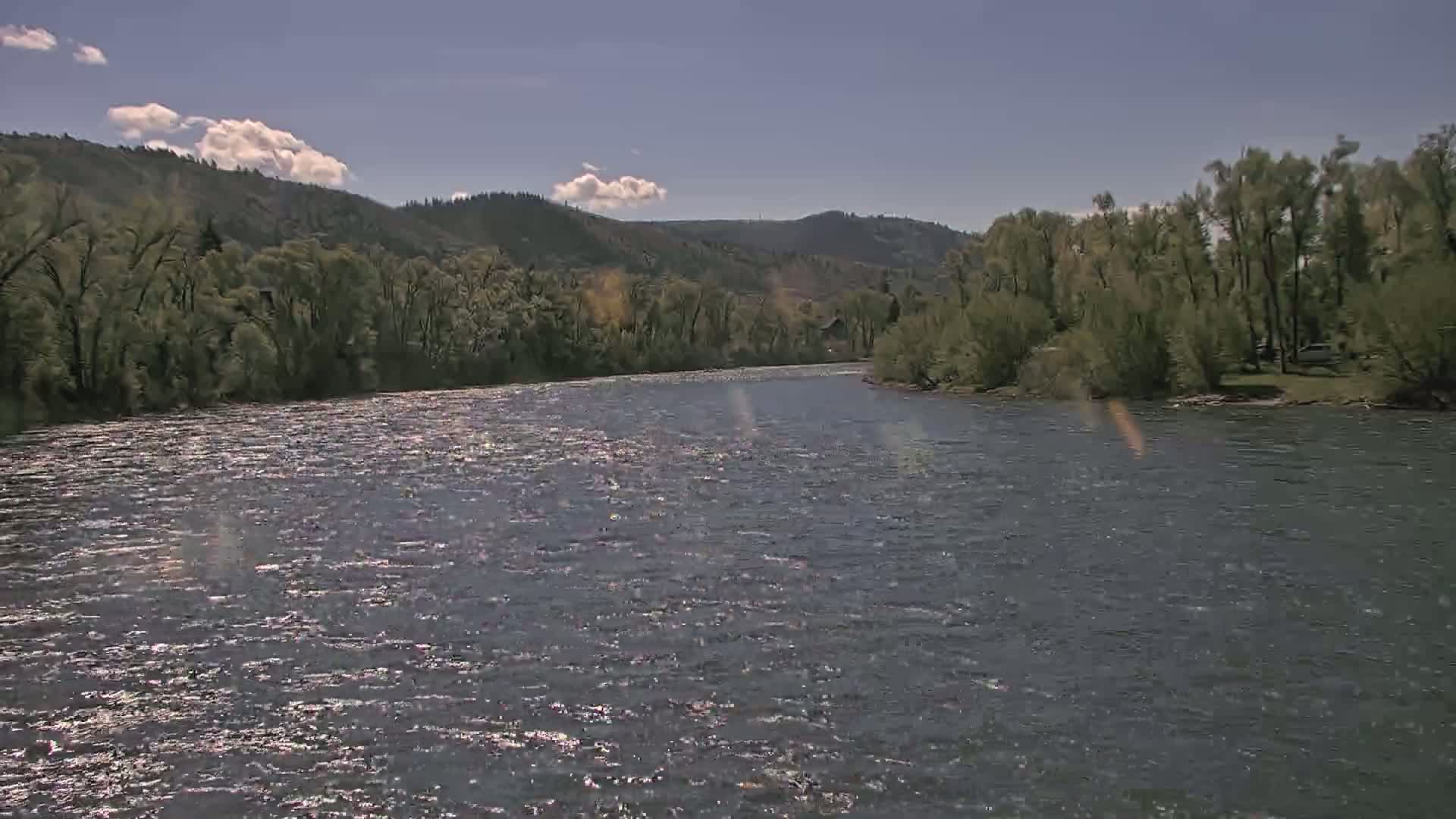A wide, gently flowing river under a partly sunny sky is bordered by lush green trees and hills.