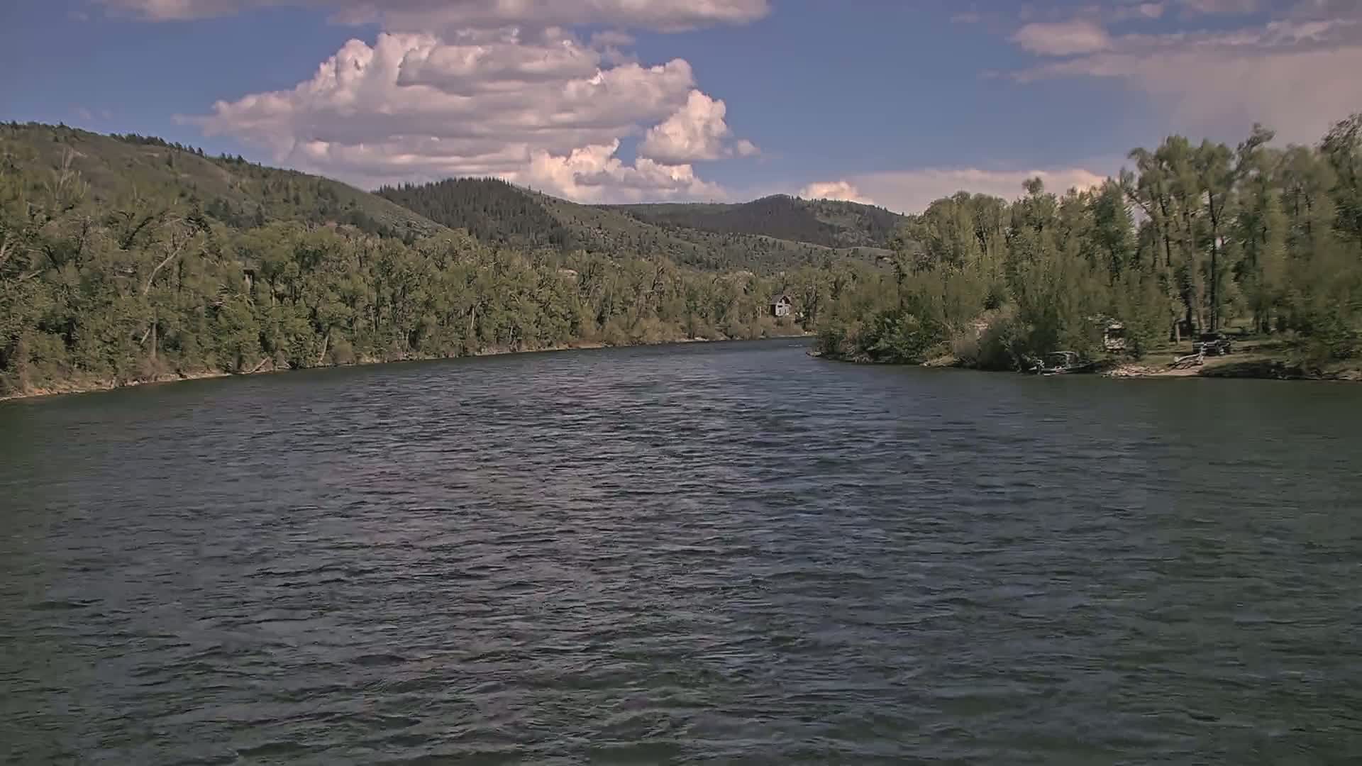 A wide river flows under a partly cloudy sky, flanked by lush green vegetation and hills.