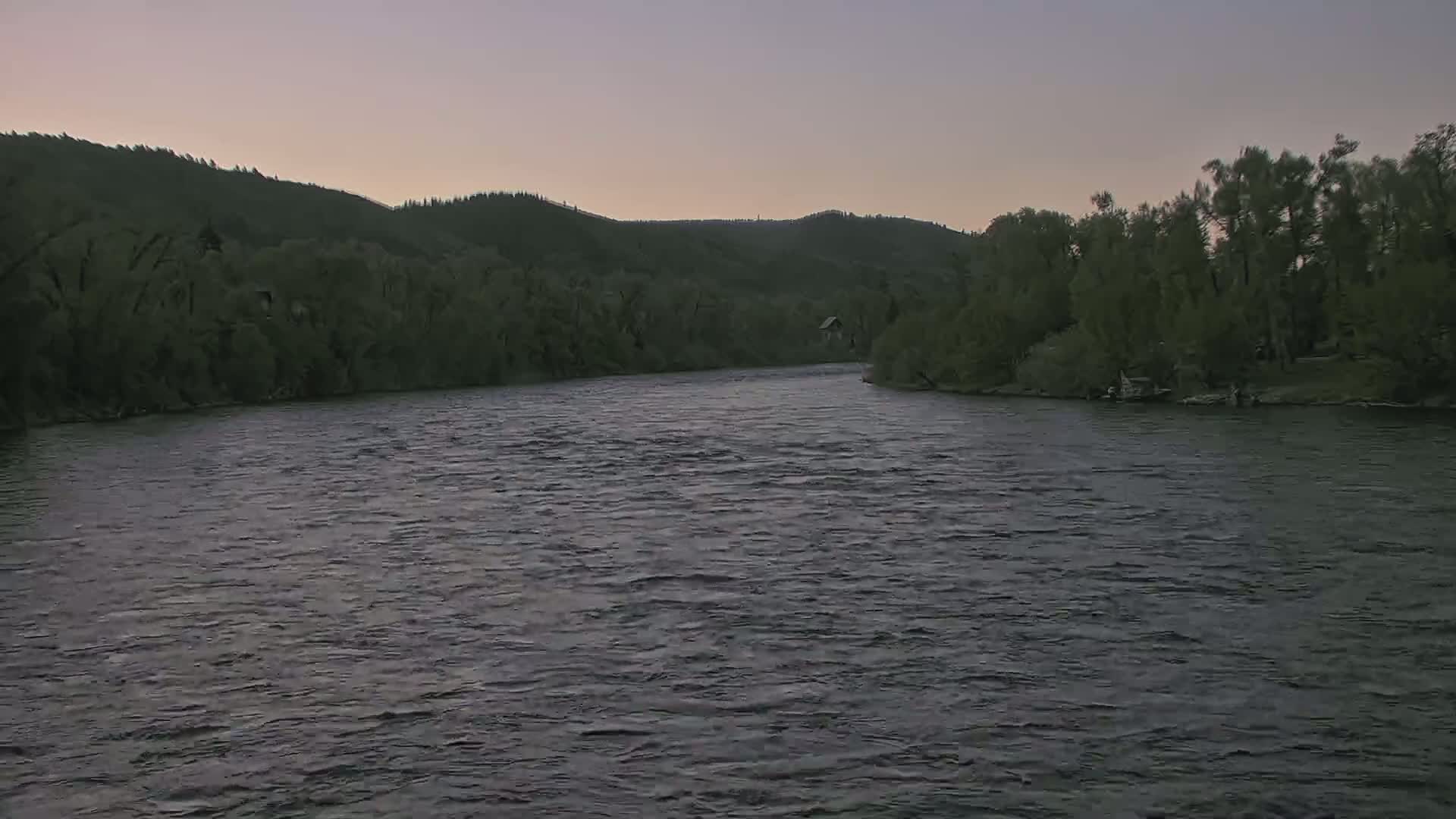 A wide, dark river flows under a dusky sky past tree-lined banks and hills.