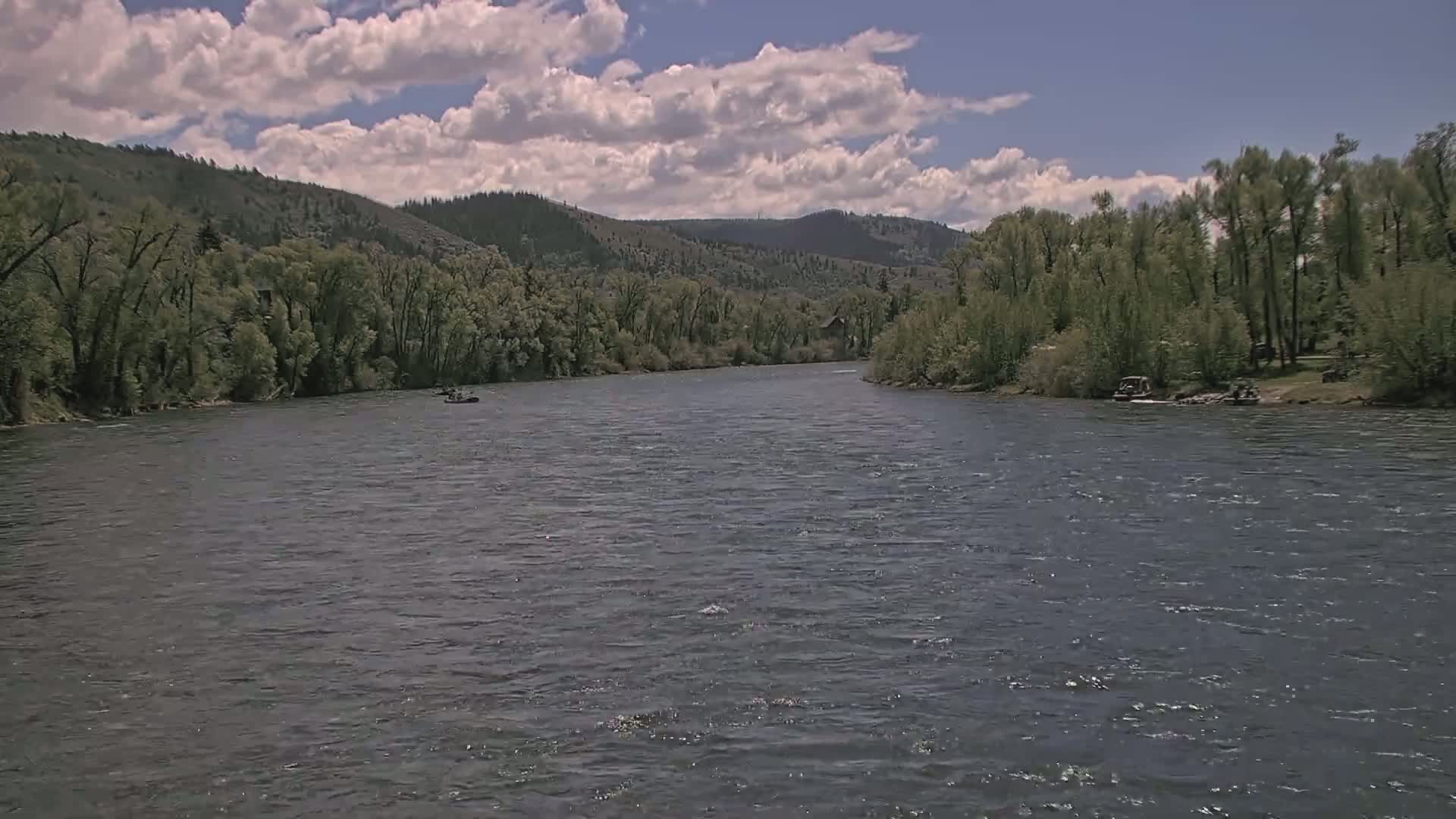 A wide, dark river flows under a partly cloudy sky, past lush green banks lined with trees and a few small boats.