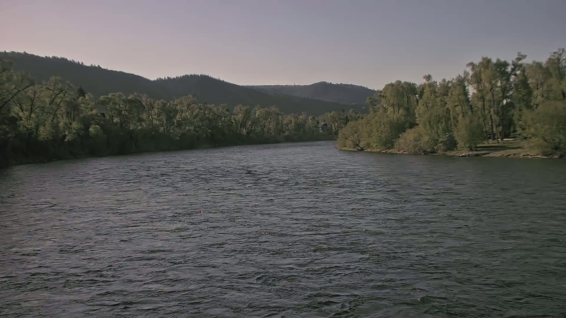 A wide, dark river flows gently under a pale sky, past tree-lined banks and a distant mountain range.