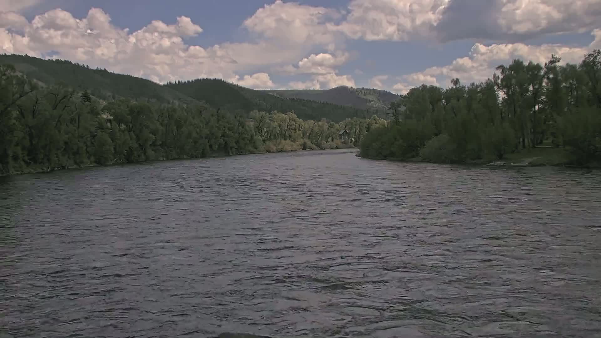 A wide river flows under a partly cloudy sky past lush green banks and forested hills.
