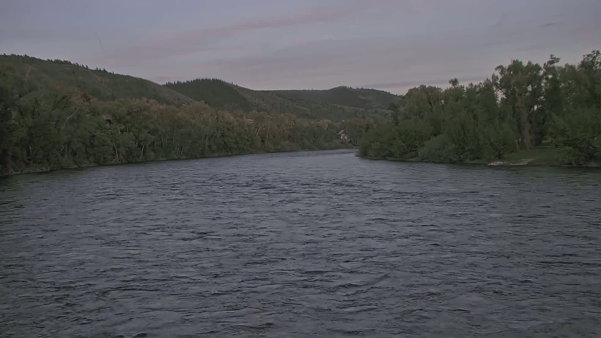 A wide, dark river flows gently past lush green hillsides under an overcast sky.