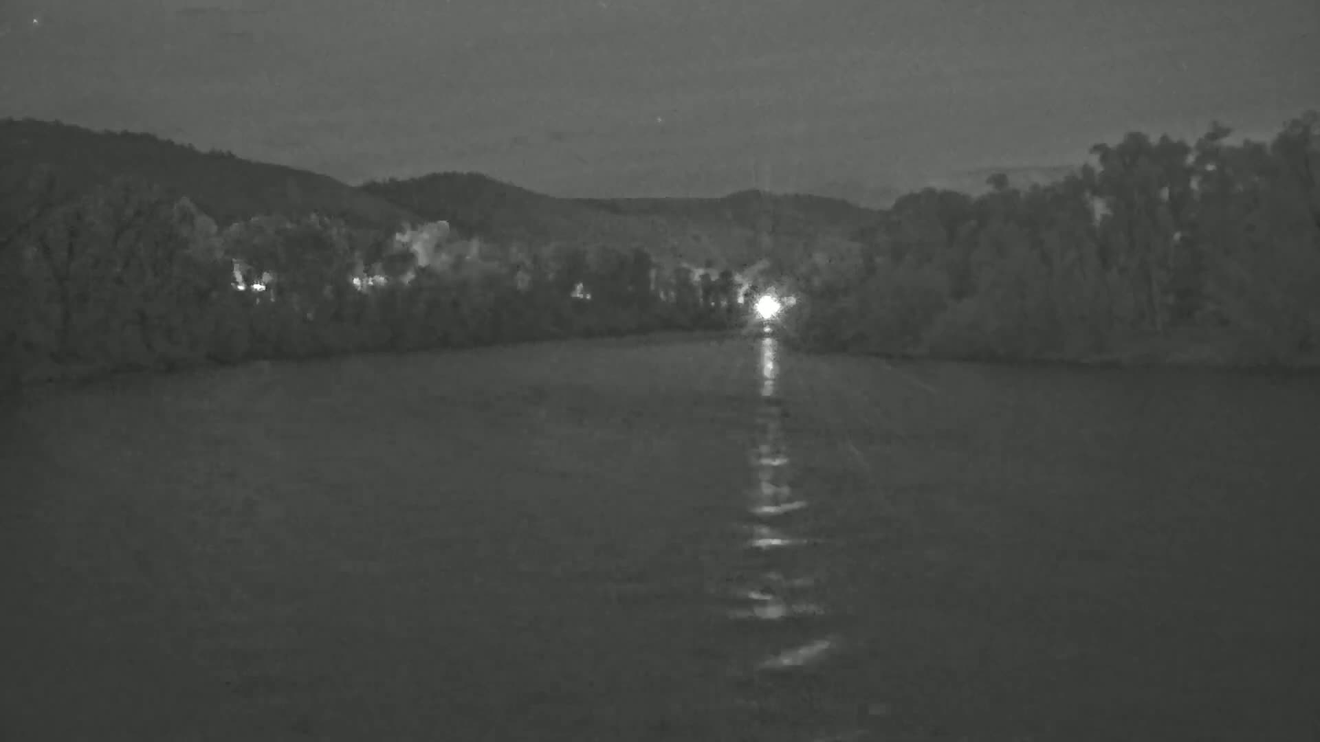 A dark, calm body of water reflects a single bright light emanating from the far bank, which is lined with trees and hills under a night sky.