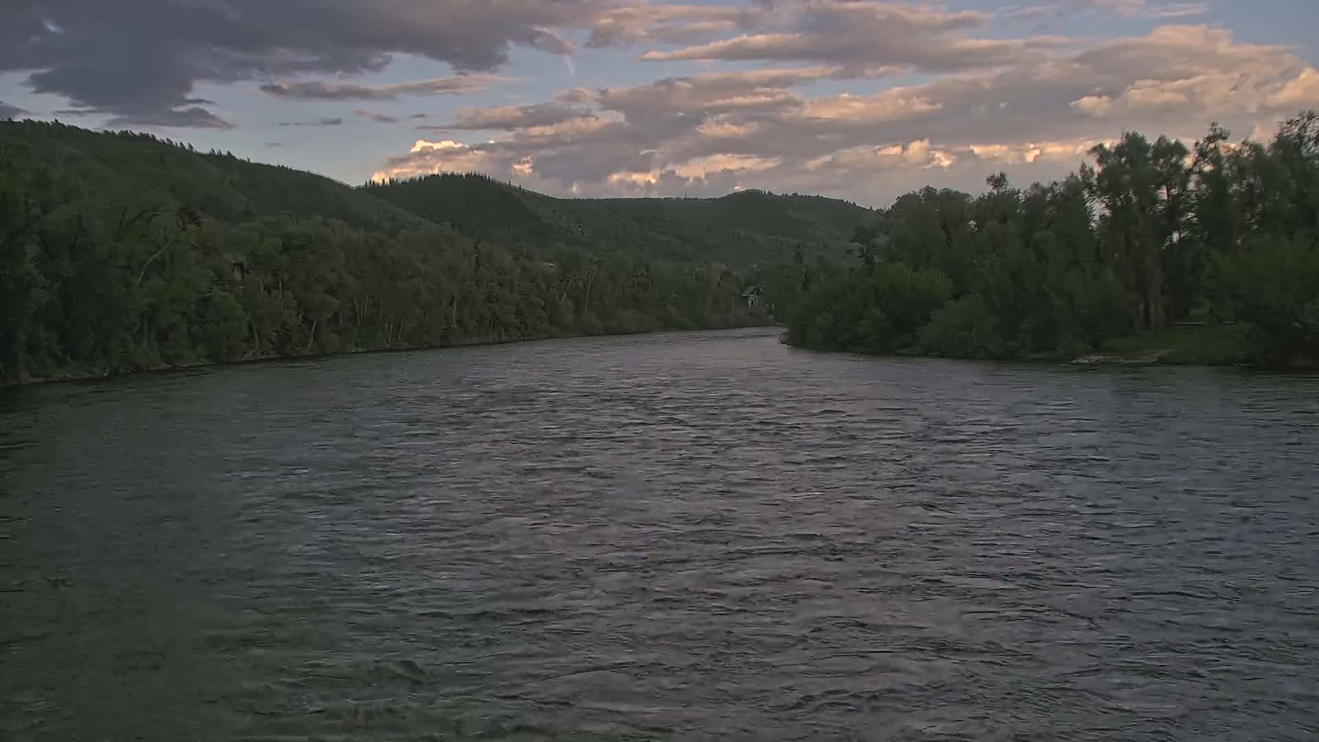 A wide, dark river flows gently under a partly cloudy, dusky sky, bordered by lush green hills and trees.