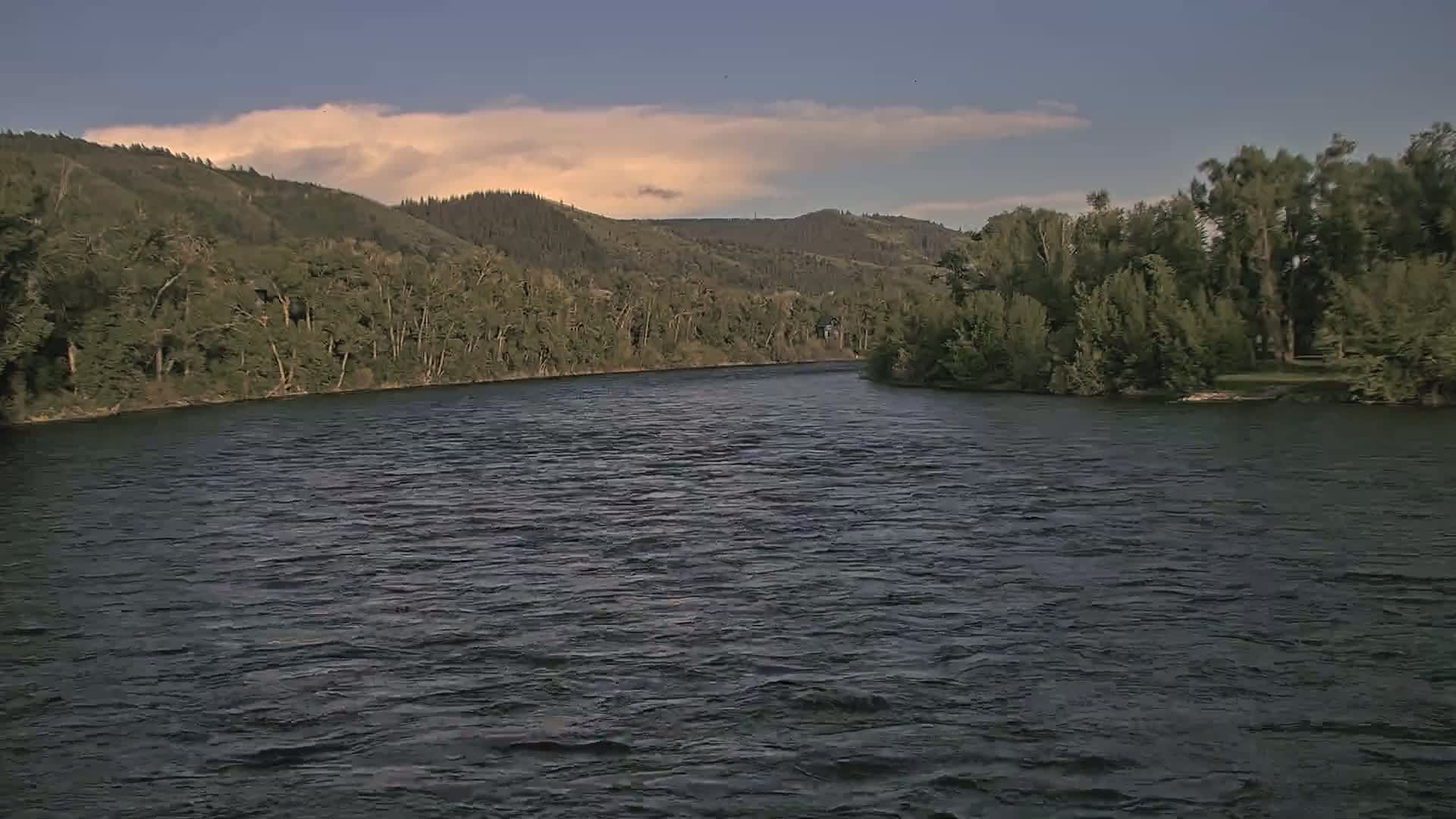 A wide, dark river flows gently under a partly cloudy sky, past lush green banks lined with trees and hills in the background.