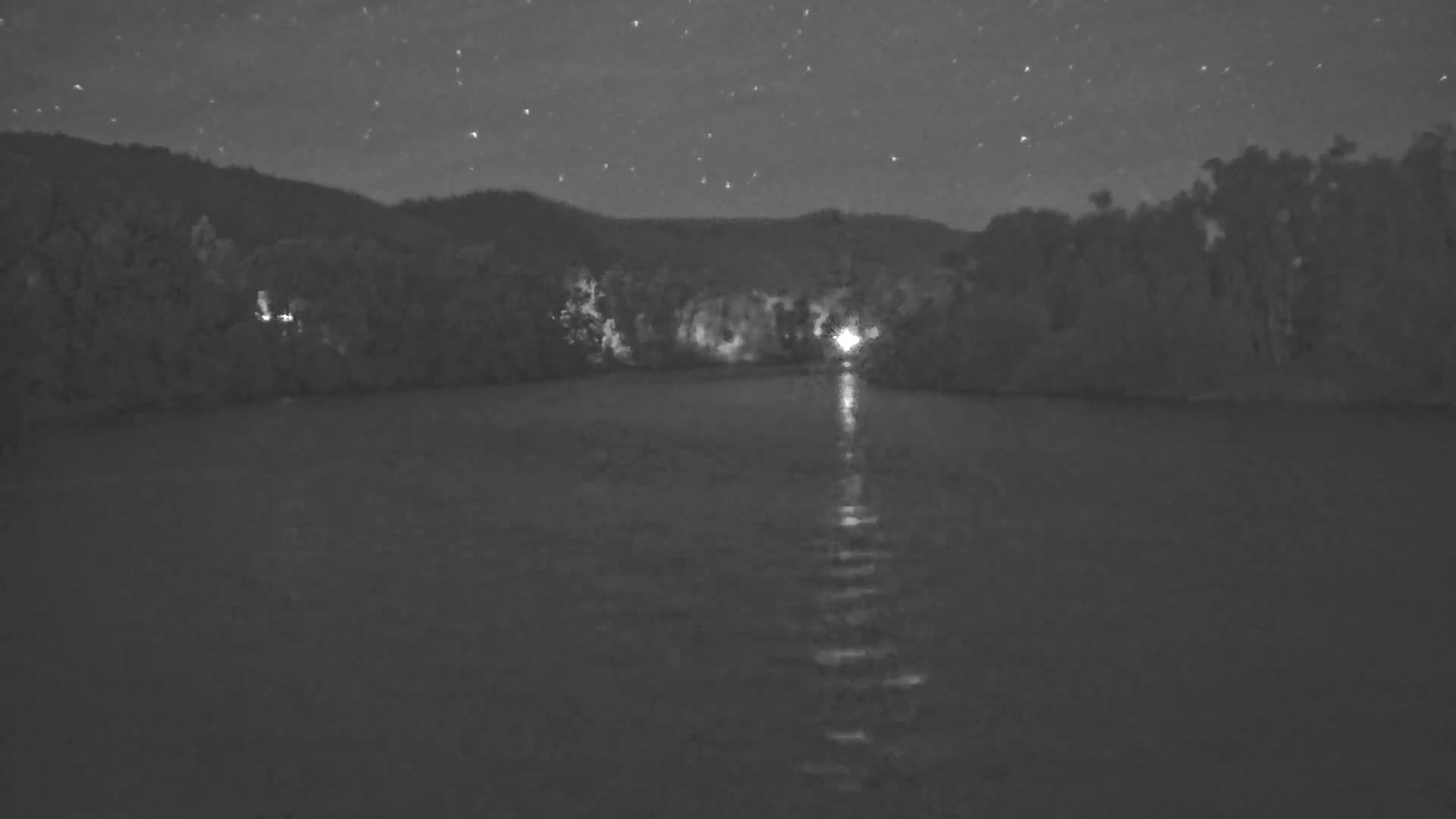 A calm body of water reflects the lights from a shoreline at night under a starry sky.