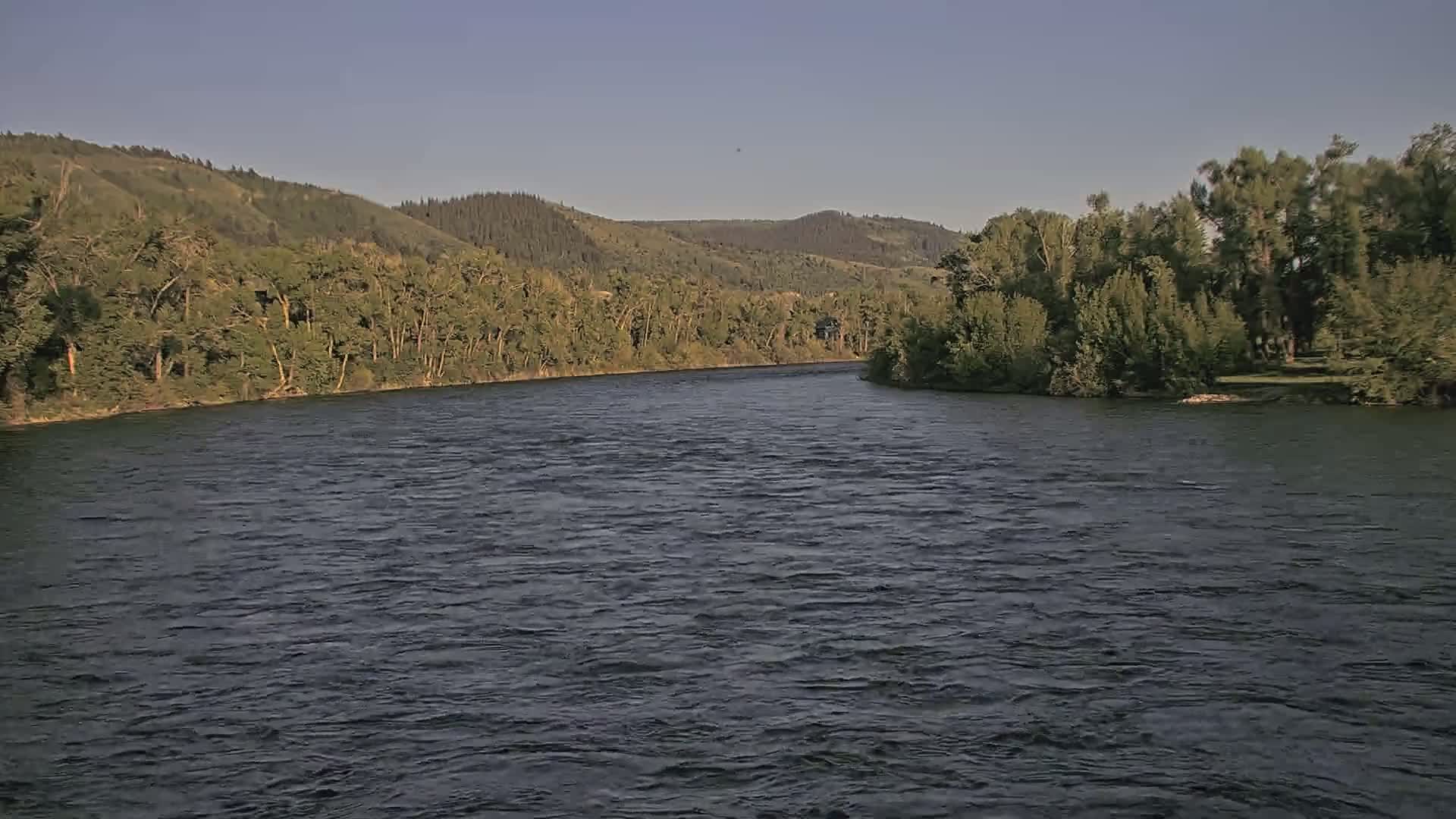 A wide, dark river flows under a clear sky past tree-lined banks and hills in the distance.
