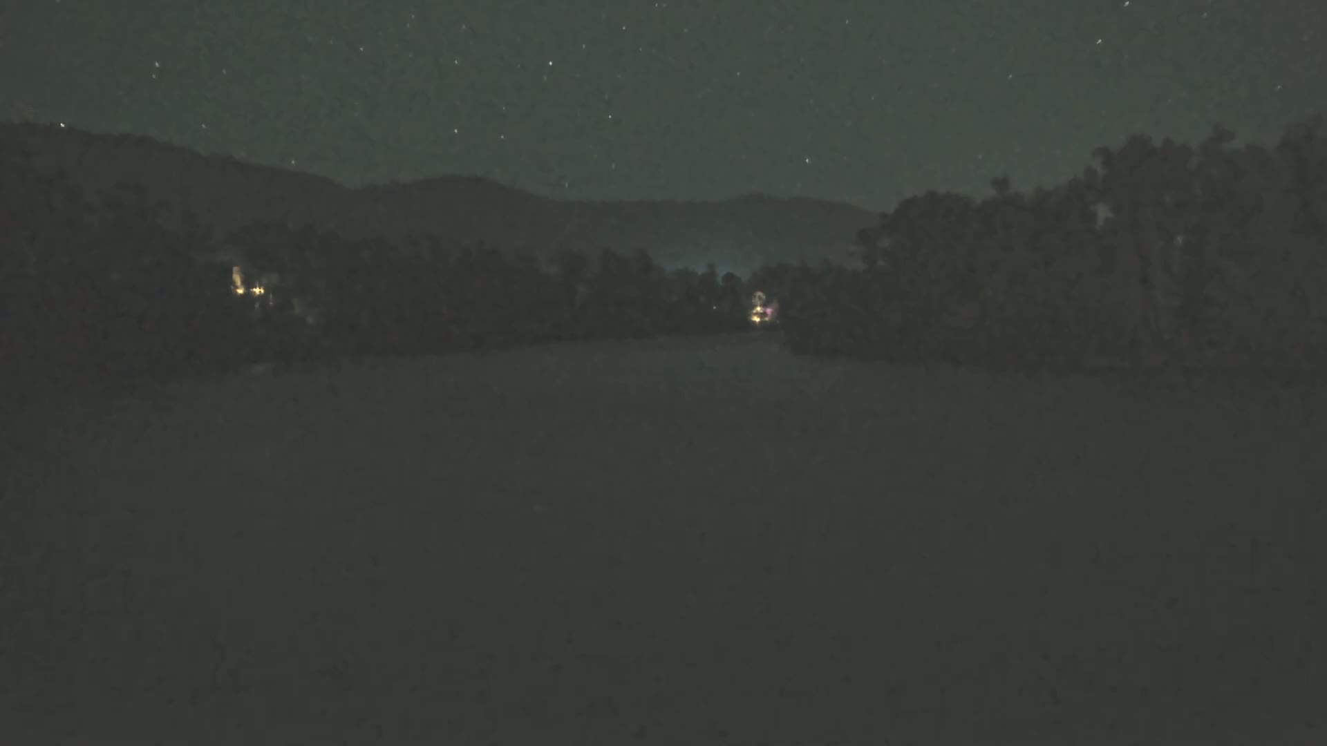 A dark, nighttime landscape shows a line of trees silhouetted against a starry sky and mountains in the distance, with a few faint lights visible near the trees.