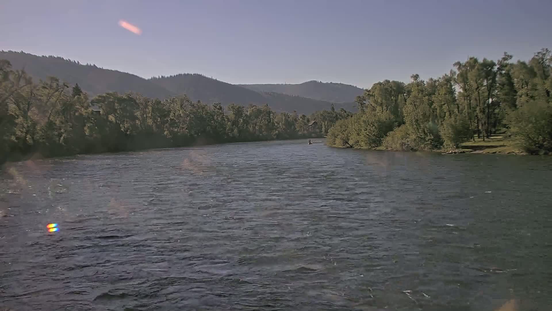 A wide, dark river flows beneath a clear sky past lush green banks and distant mountains.