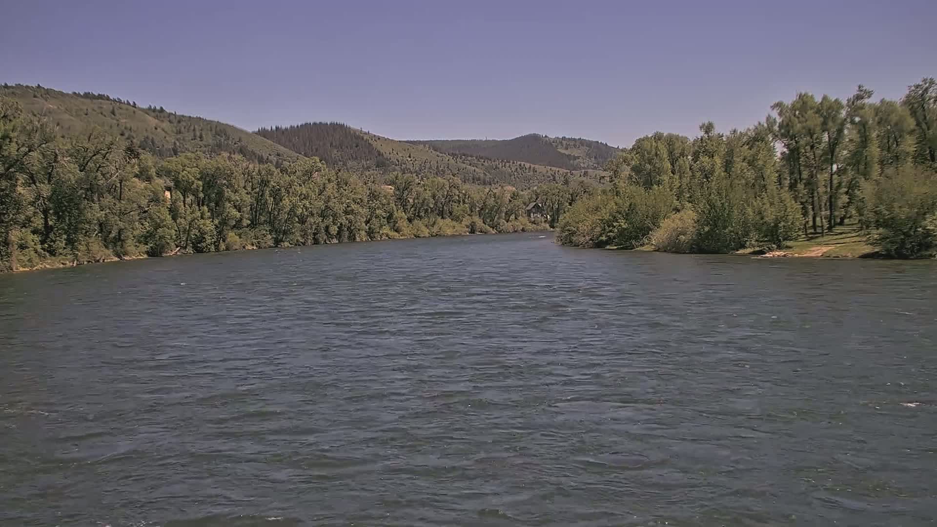 A wide, dark river flows between lush green hills under a clear, sunny sky.