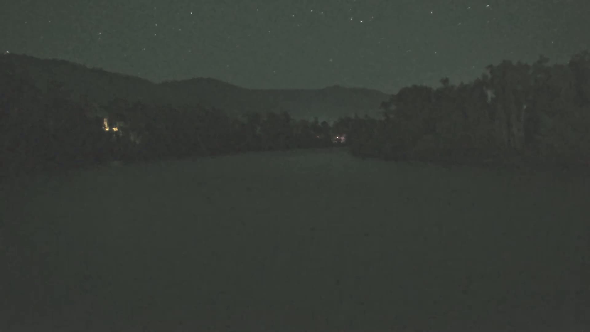 A dark, flat landscape under a starry night sky shows a line of trees and distant mountains.