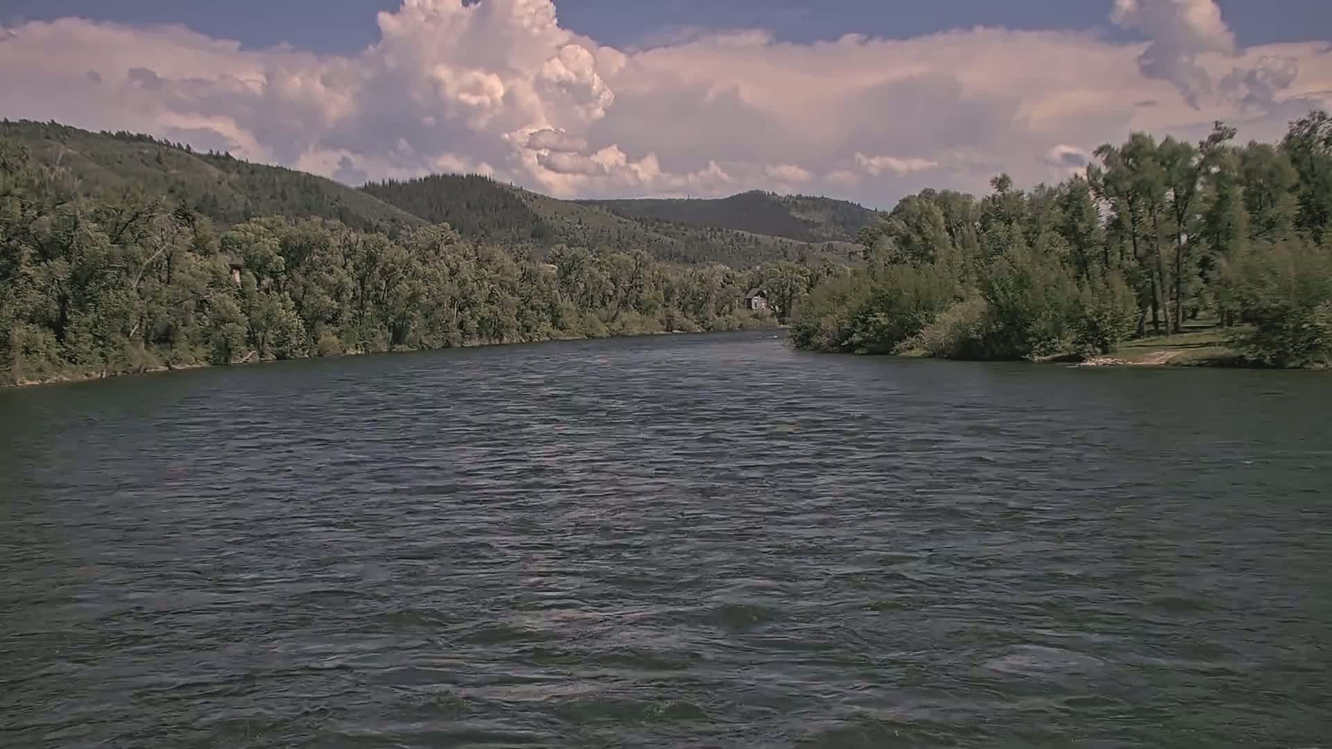A wide, dark river flows beneath a partly cloudy sky past lush green banks lined with trees and hills in the distance.