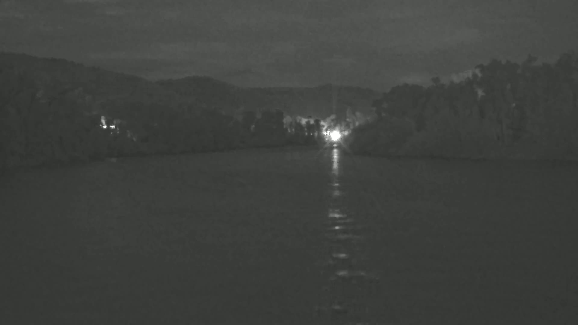 A nighttime view of a body of water reflecting the light of a distant source, with a dark shoreline lined with trees in the background under a cloudy sky.