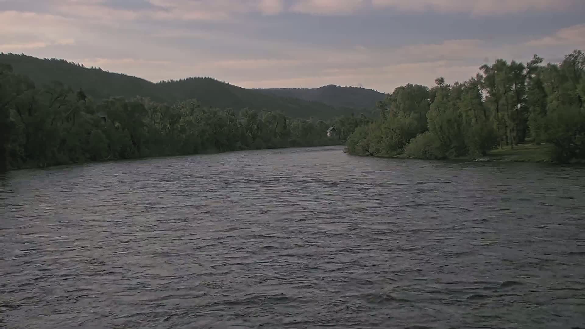 A wide, somewhat choppy river flows past lush green trees and hills under a cloudy sky.