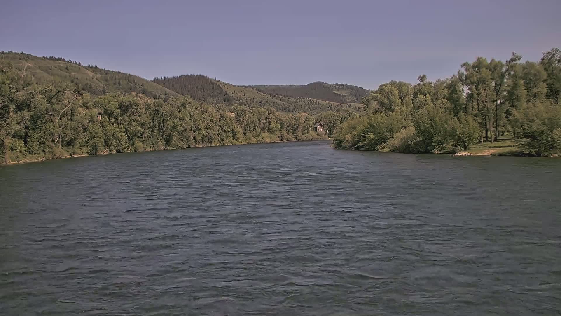 A wide, dark river flows under a clear sky past forested hillsides on a sunny day.