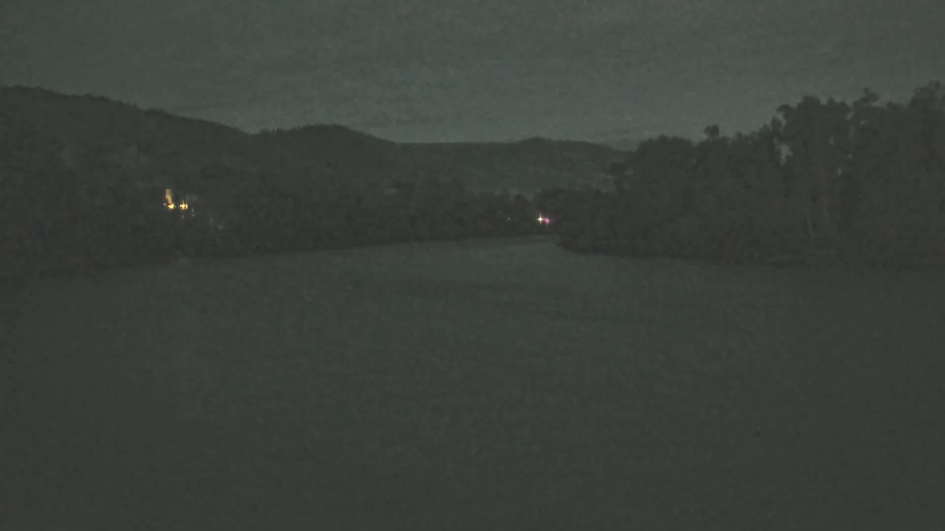 A dark, nighttime scene shows a calm body of water reflecting a dark sky, with dimly lit trees and hills in the background.