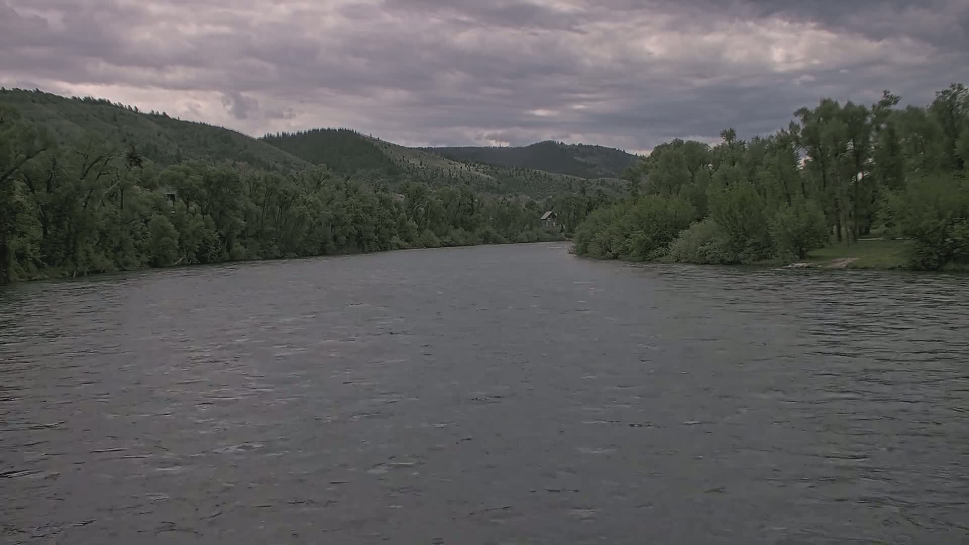 A wide, grey river flows under an overcast sky past lush green hillsides lined with trees.
