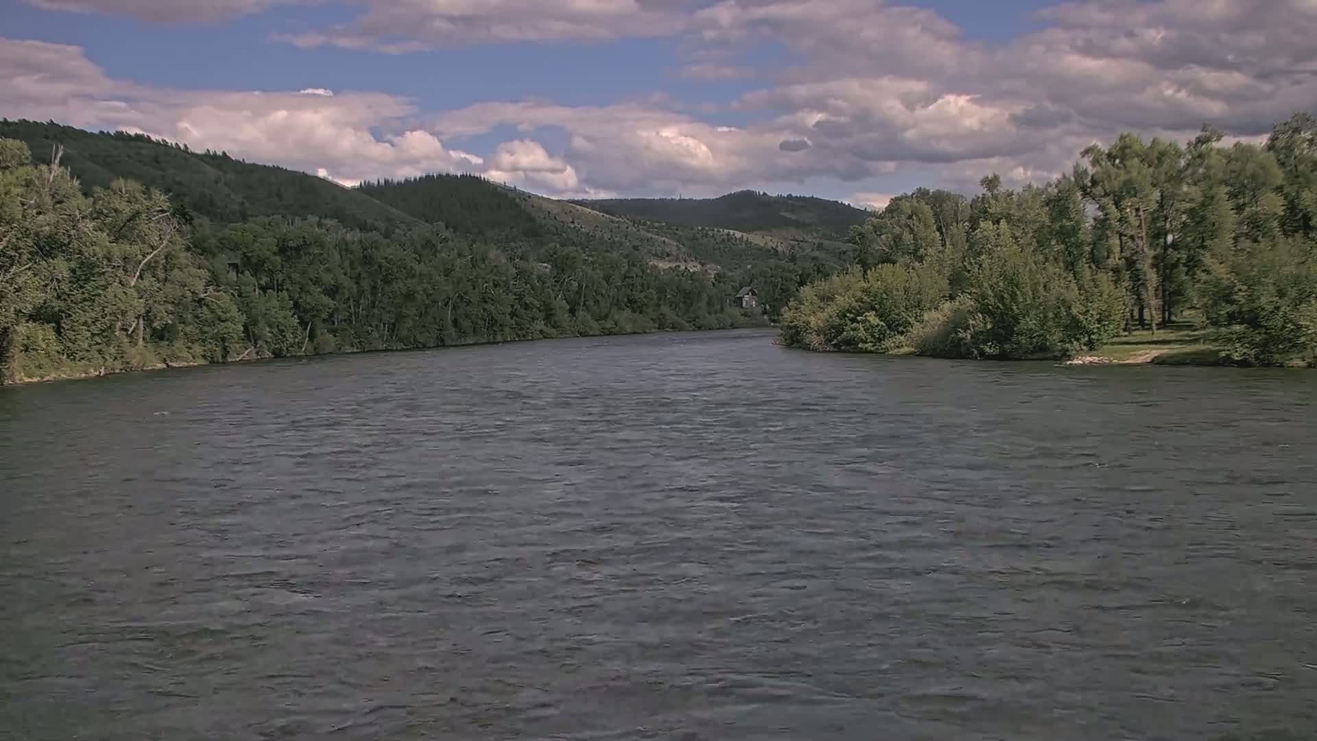 A wide, calm river flows between lush green banks under a partly cloudy sky.