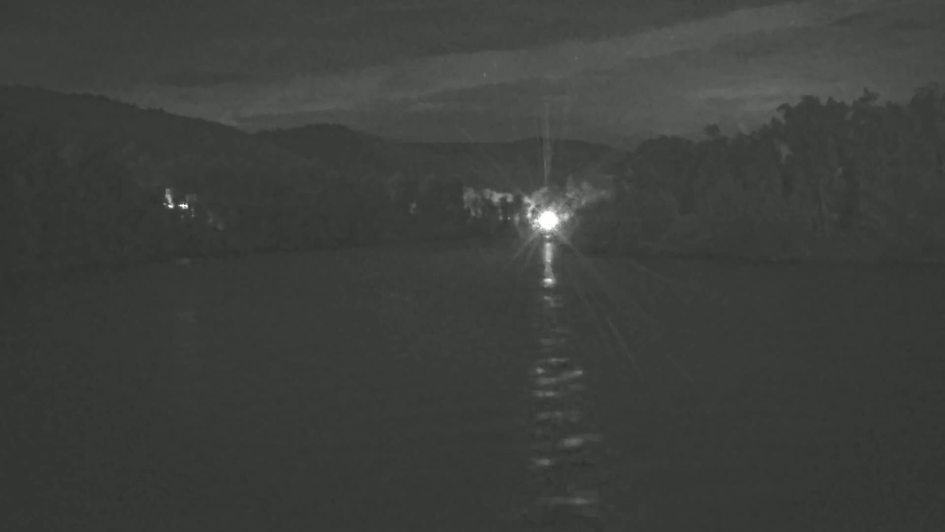A bright light shines from a distant vessel on dark water under a night sky, with dark hills and trees in the background.