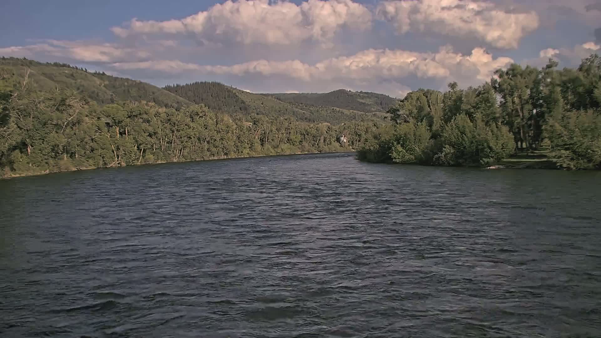 A wide, dark river flows under a partly cloudy sky, bordered by lush green trees and rolling hills.
