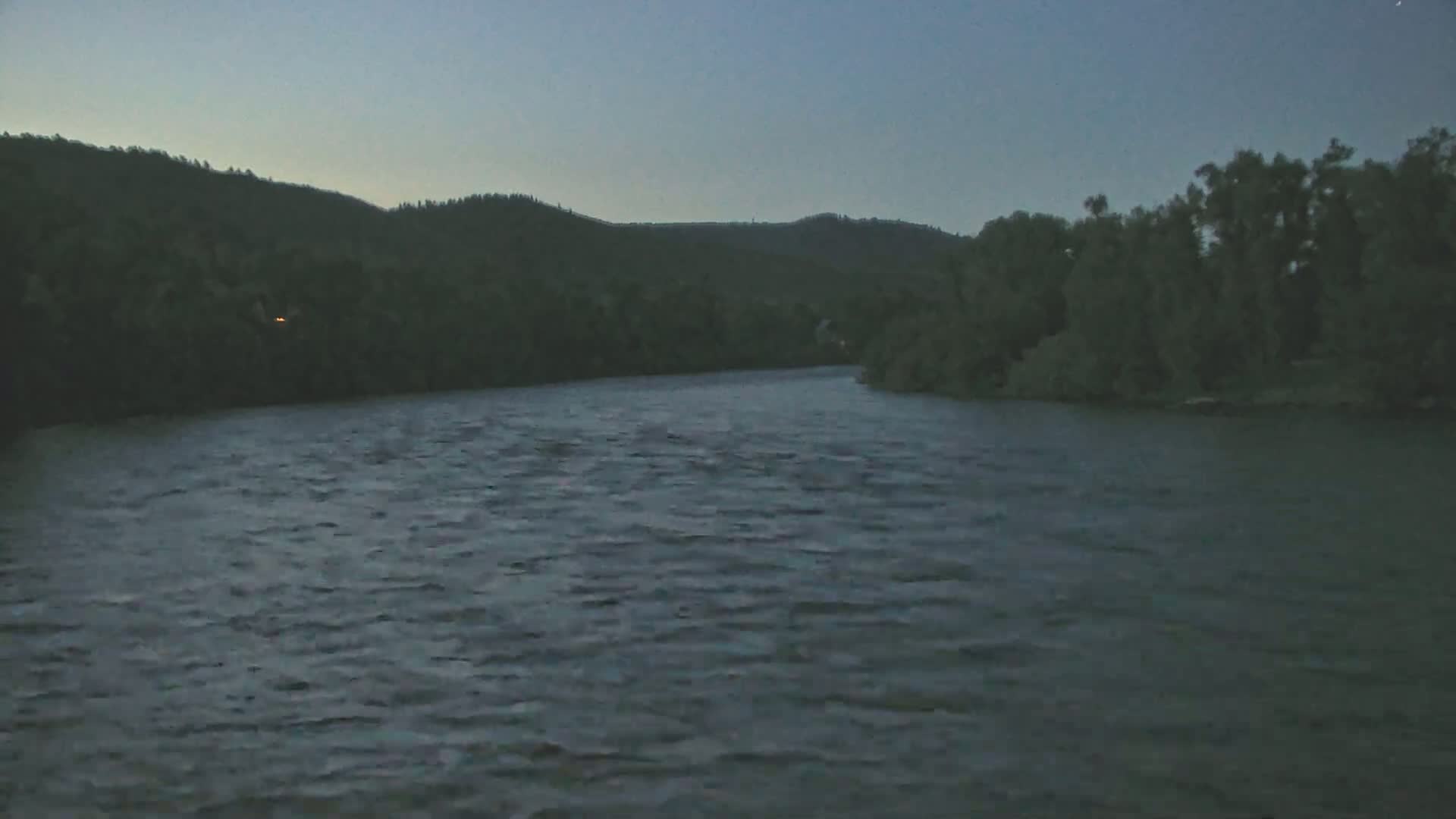 A dark, gently rippling river flows beneath a twilight sky, past a treeline and hills in the distance.