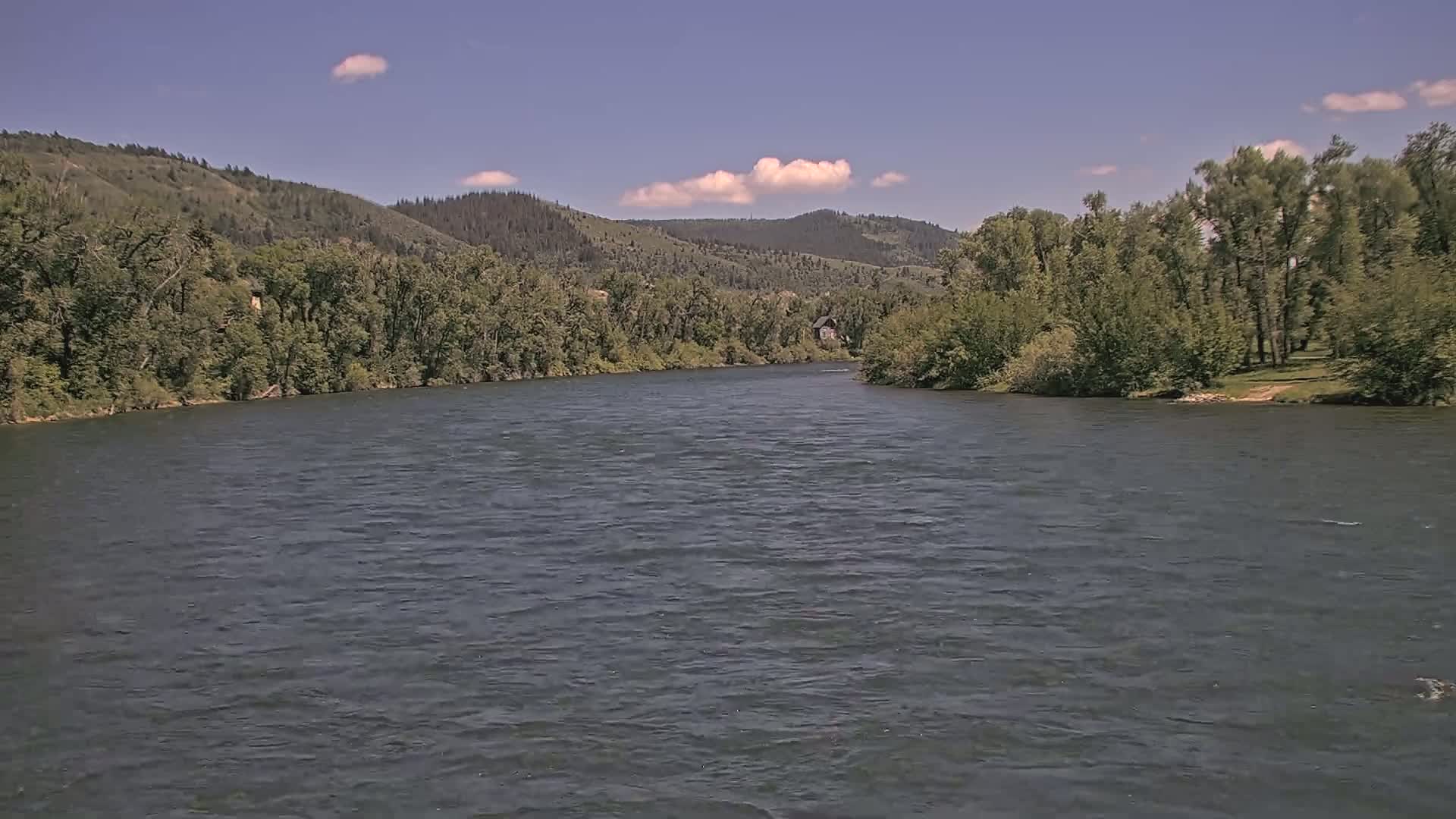 A wide, dark river flows under a mostly clear blue sky with a few small cumulus clouds, flanked by lush green trees and hills.