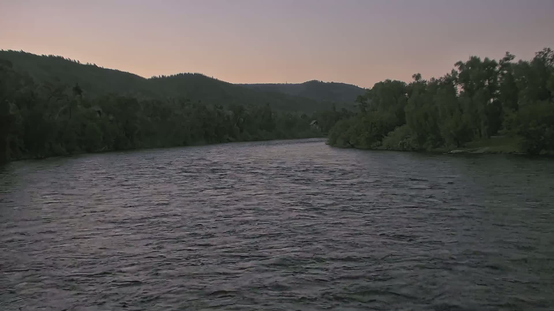A wide, dark river flows gently under a dusky sky past tree-lined banks.