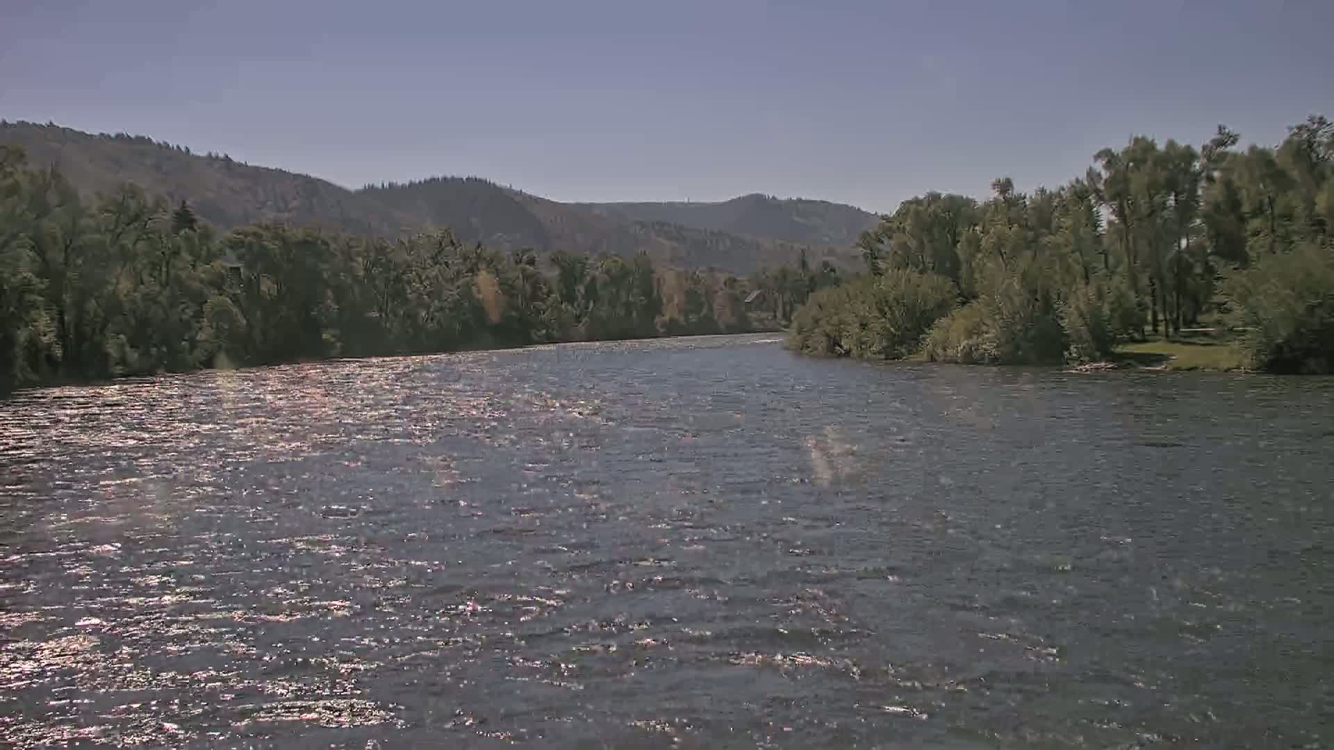 A wide, gently flowing river reflects sunlight under a clear blue sky, bordered by lush green trees and distant hills.