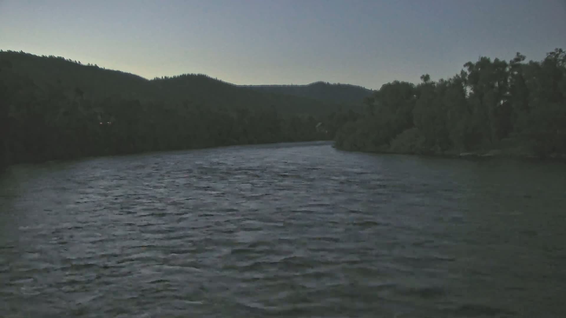 A wide, dark river flows under a dusky sky past tree-lined banks and a dark mountain range in the distance.