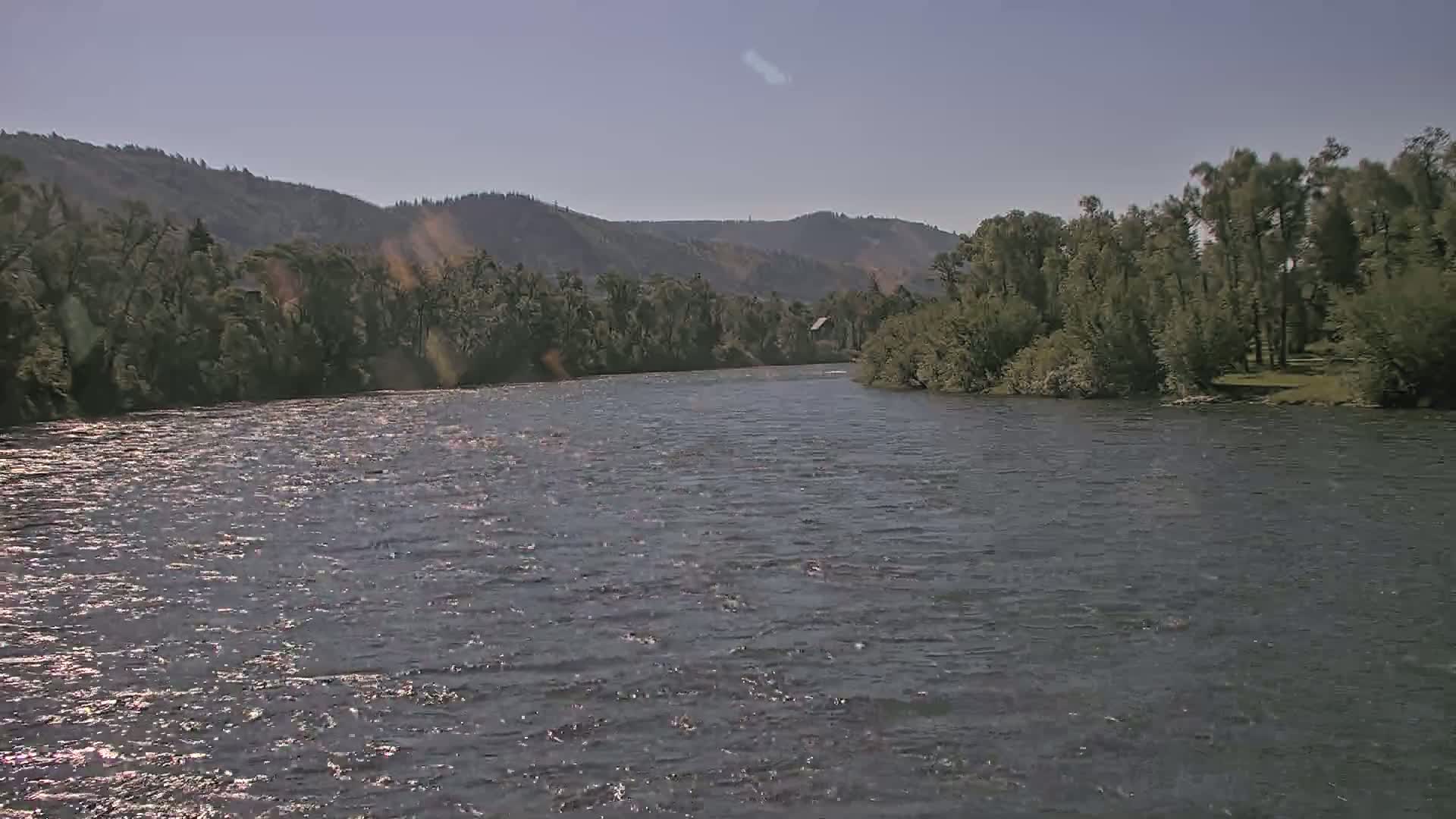 A wide, gently flowing river is seen under a clear sky, bordered by lush green vegetation and distant hills.