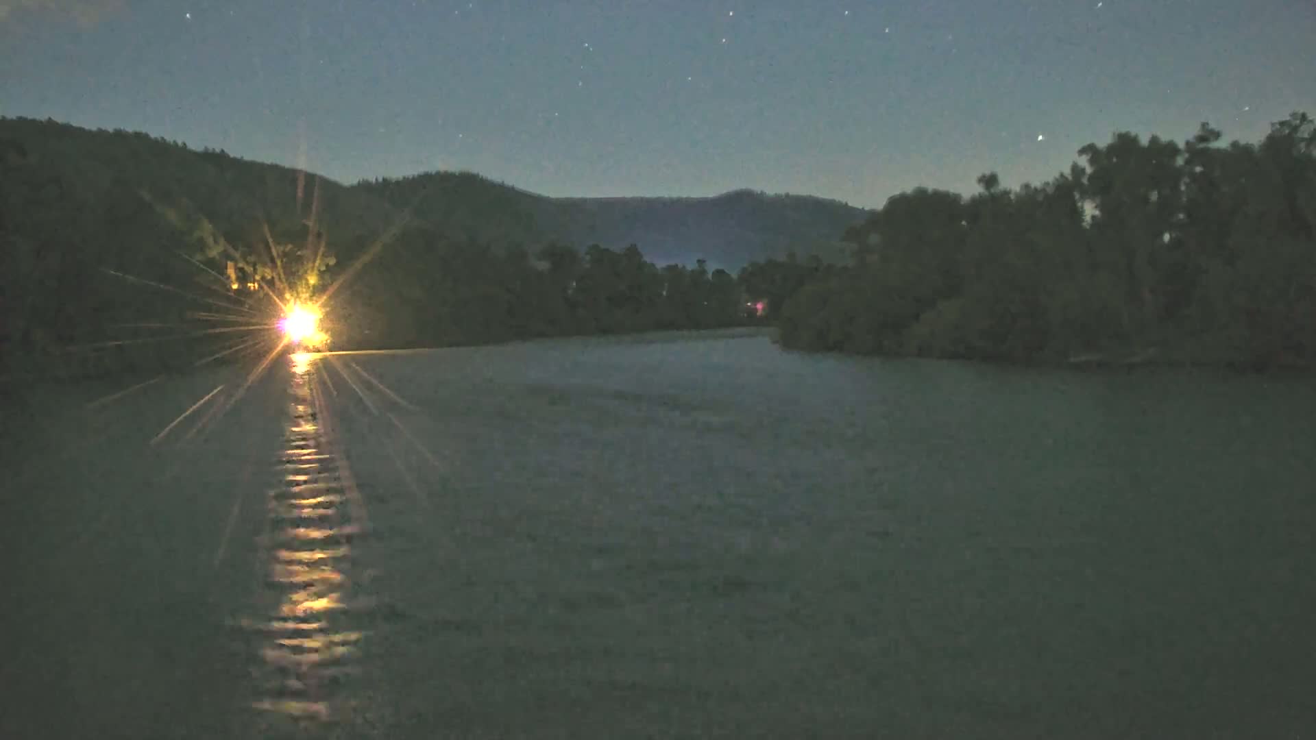 A bright light on a body of water at night, surrounded by dark hills and trees under a starry sky.