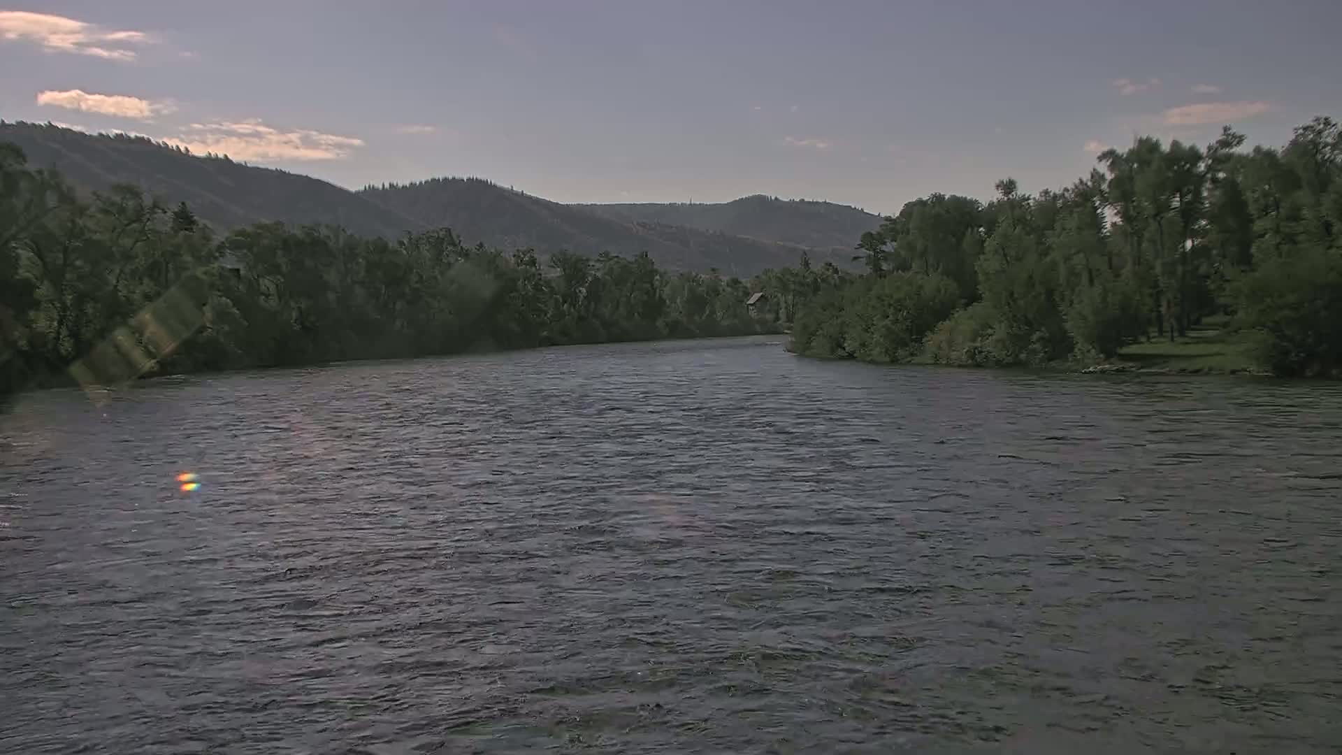 A wide, gently flowing river under a partly cloudy sky is bordered by lush green trees and hills.