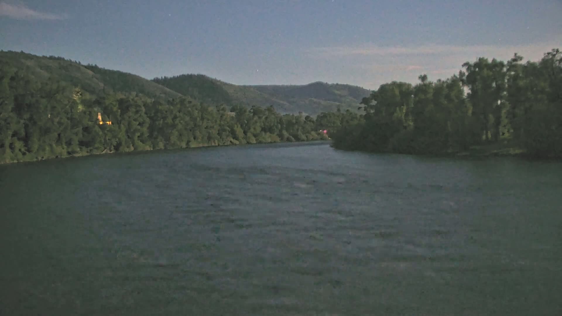 A dark river flows under a twilight sky past lush green hillsides covered in trees.