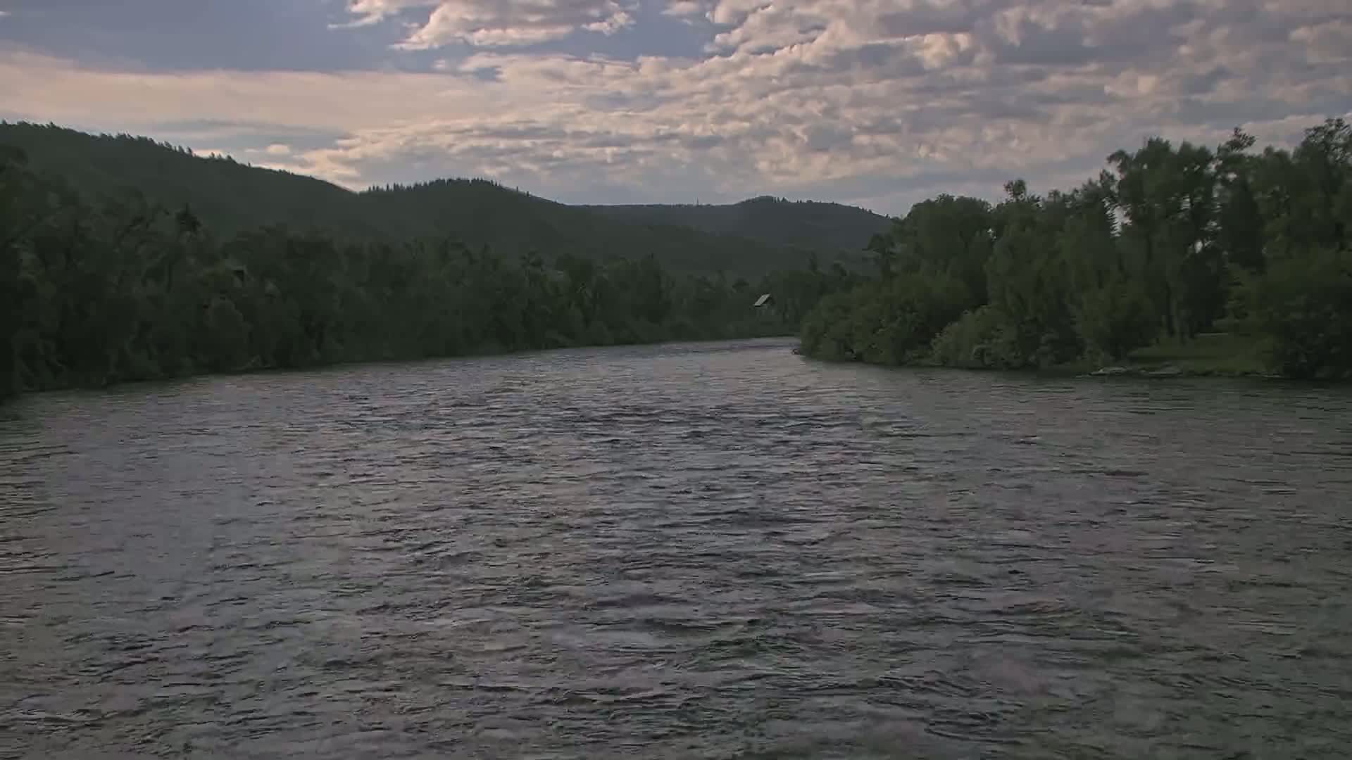 A wide, gently flowing river is surrounded by lush green trees and hills under a partly cloudy sky.