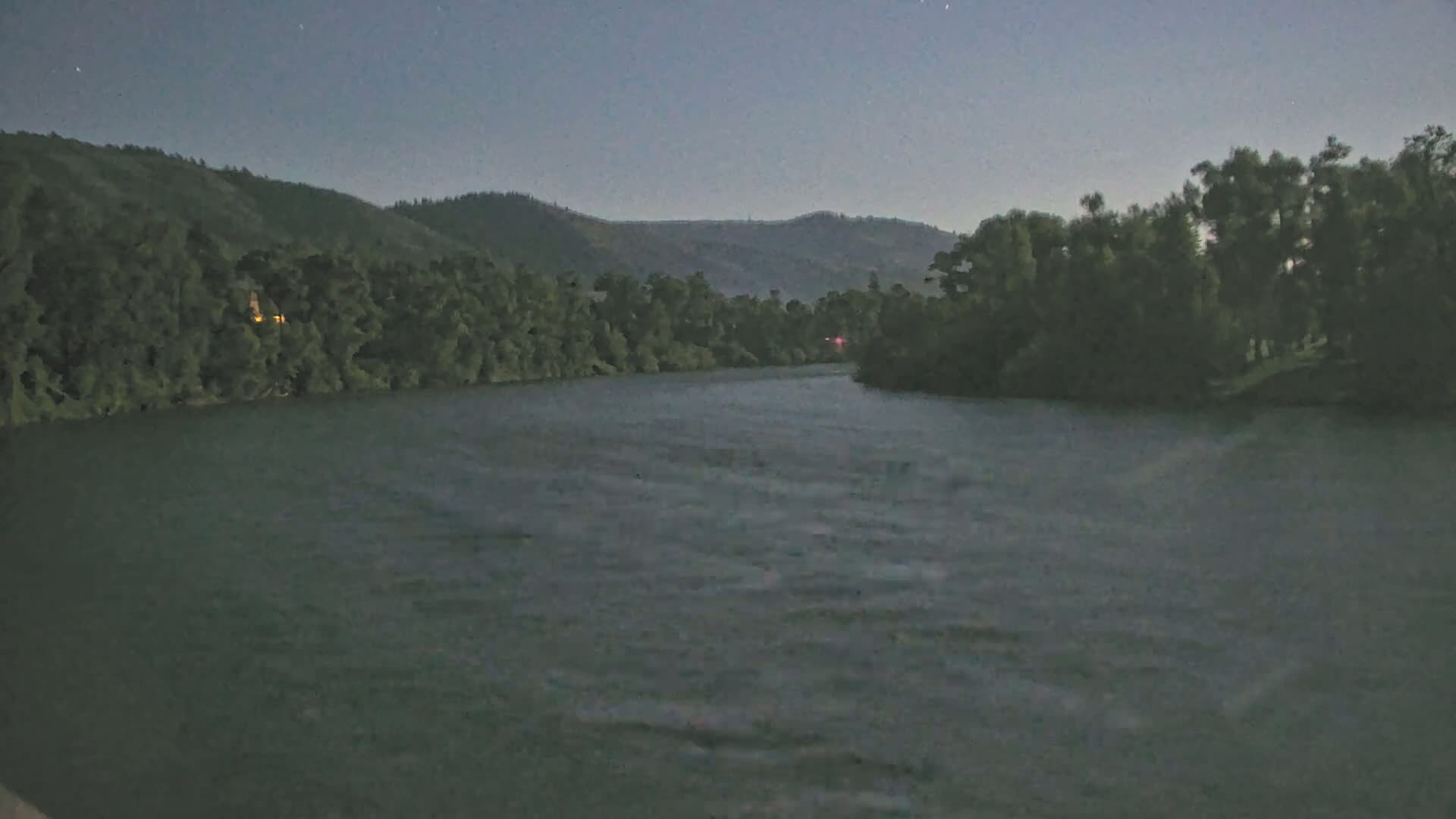A dark, calm river flows under a twilight sky past tree-lined banks and distant hills.