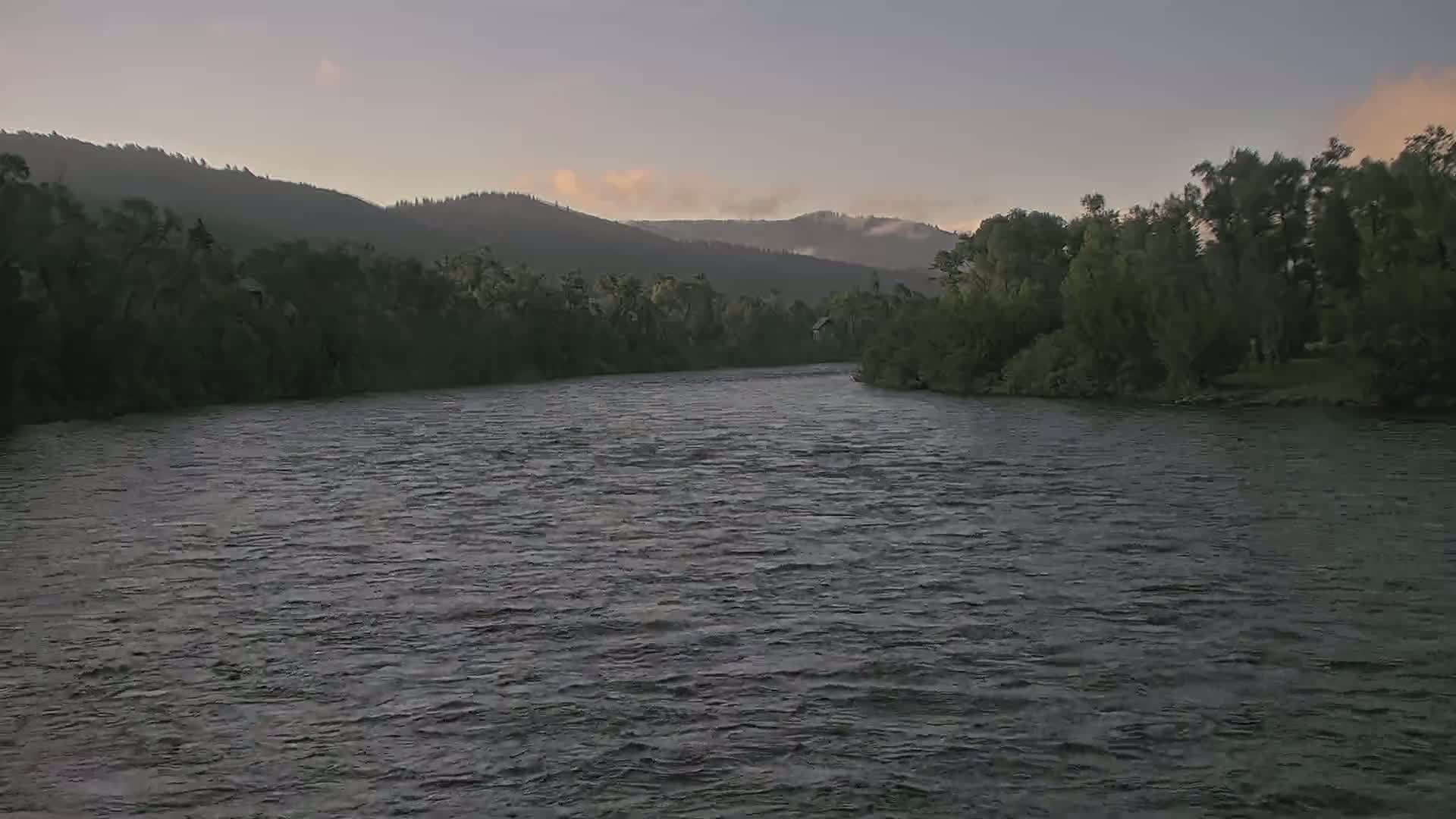 A wide, gently flowing river curves through a landscape of dark green trees and softly lit, distant mountains under a pale, dusky sky.